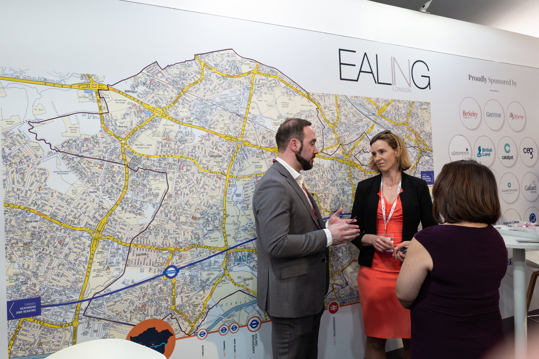 Three people having a conversation near a large London street map displayed on a wall at an event, with sponsor logos on the right side.