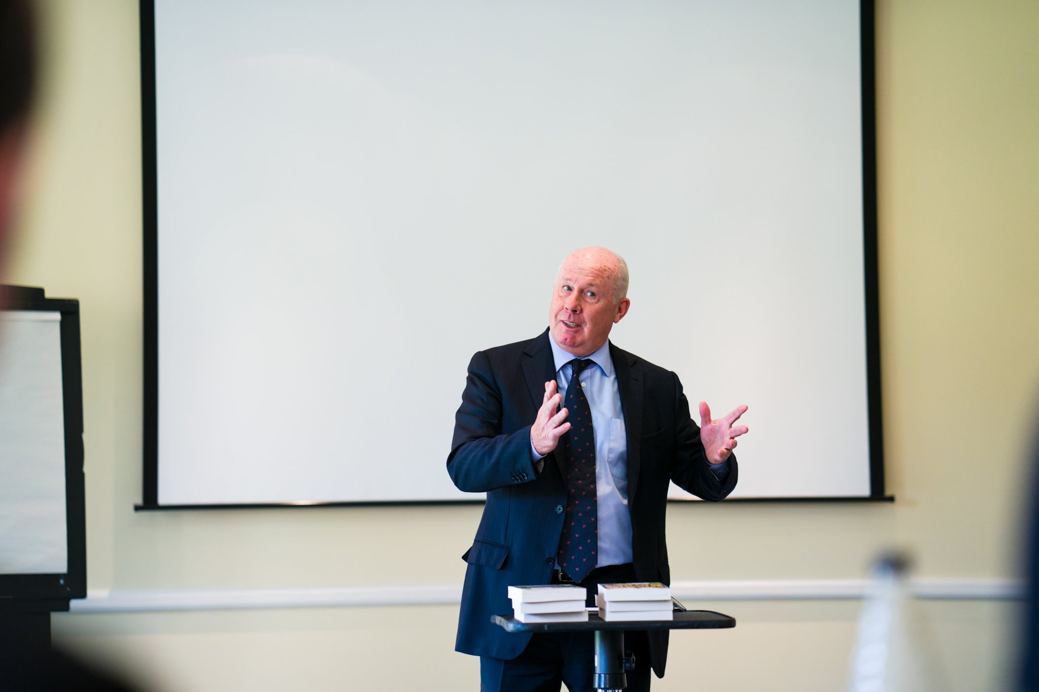 A man in a dark suit and tie is standing in front of a whiteboard, giving a presentation during a classroom or seminar. He is gesturing with his hands and has a few stacks of books on a small table in front of him.