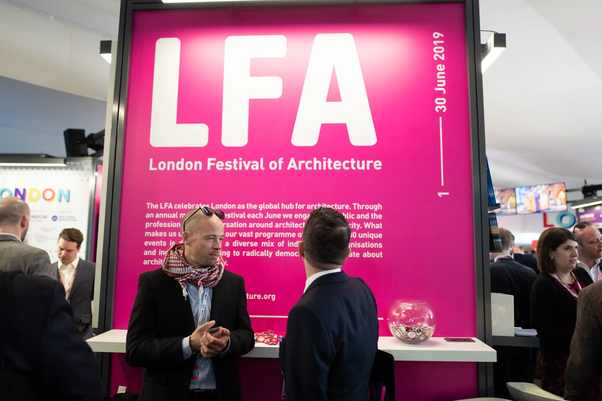 People at the London Festival of Architecture booth, with a large pink sign and text about the event.