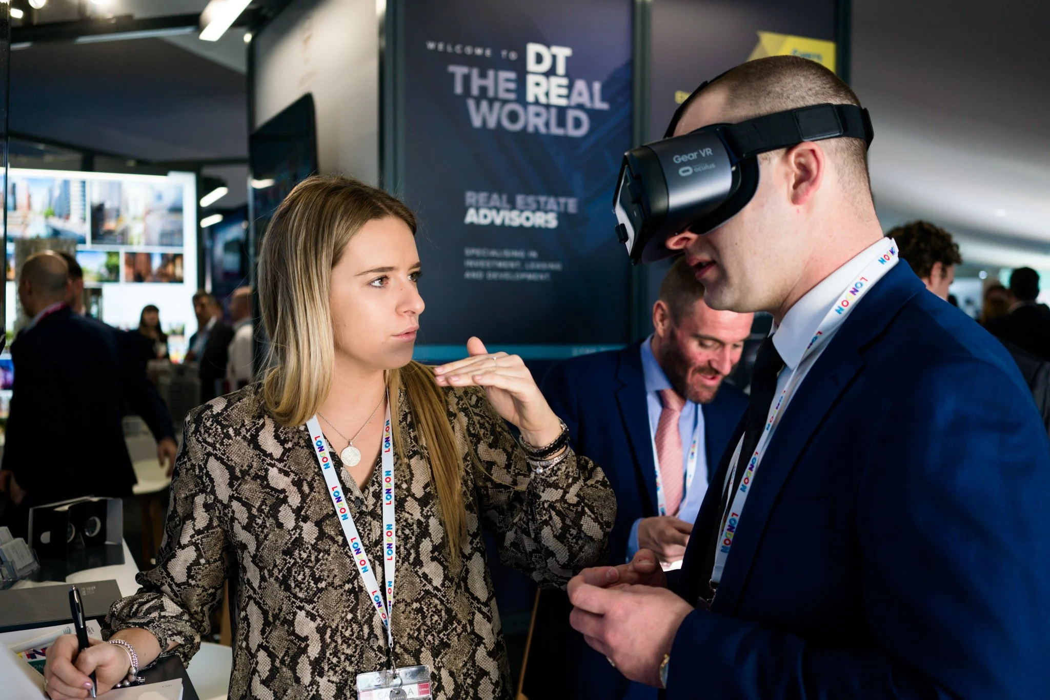 A young woman and a man wearing a virtual reality headset are engaged in a conversation at a professional event. The woman gestures with her hand while holding a pen, and the man is dressed in a suit with a lanyard around his neck.