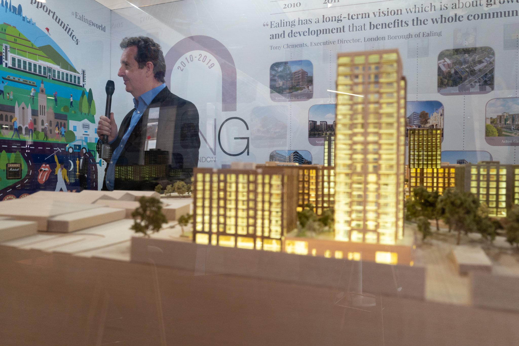 Reflective display of a man in a suit holding a microphone, with a cityscape model of illuminated buildings in the foreground, and an informational poster about urban development in the background.