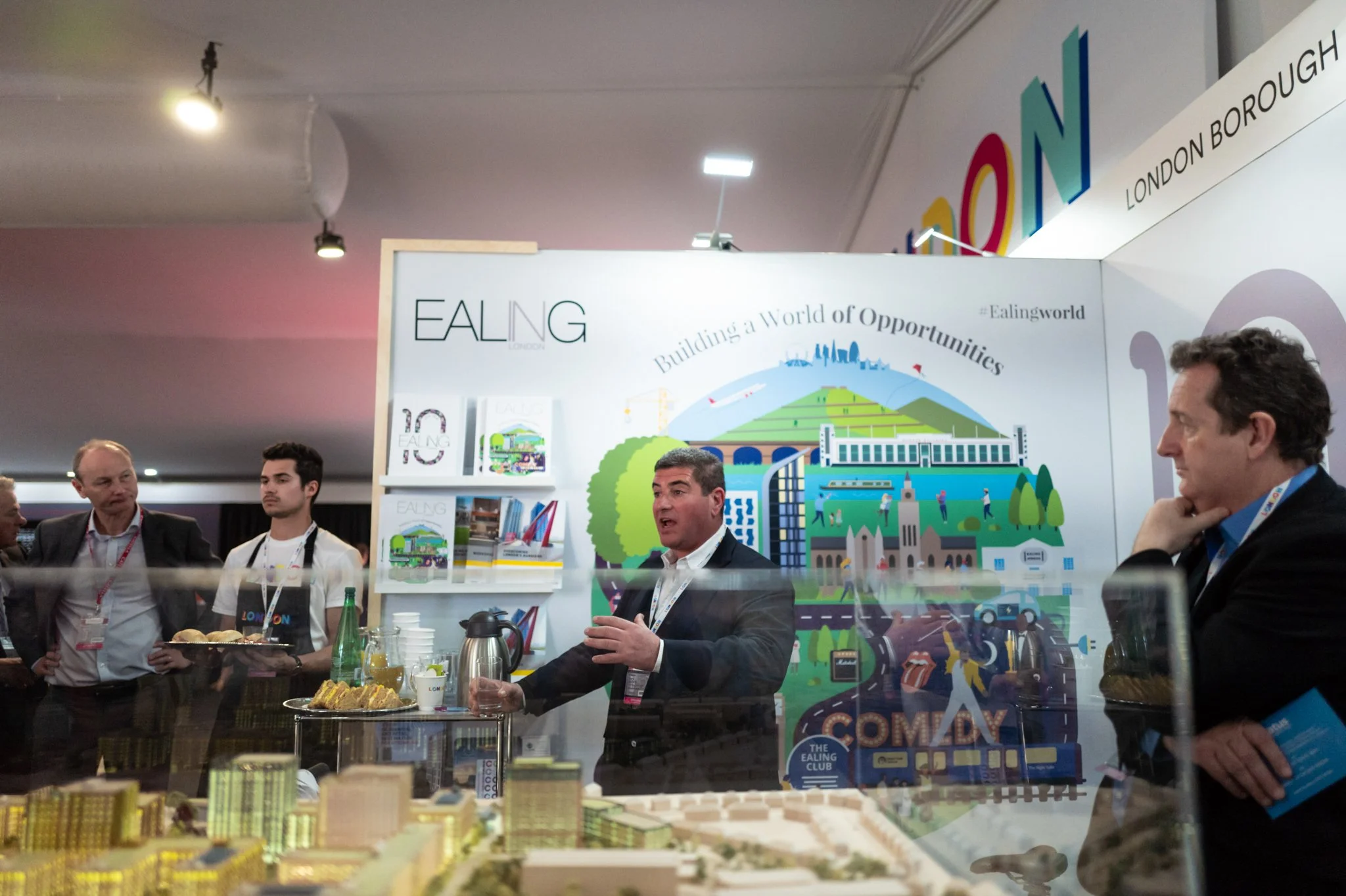 Group of people at a promotional event for Ealing in London, with one man speaking and others listening, behind a display with promotional materials and a colorful illustration of London.