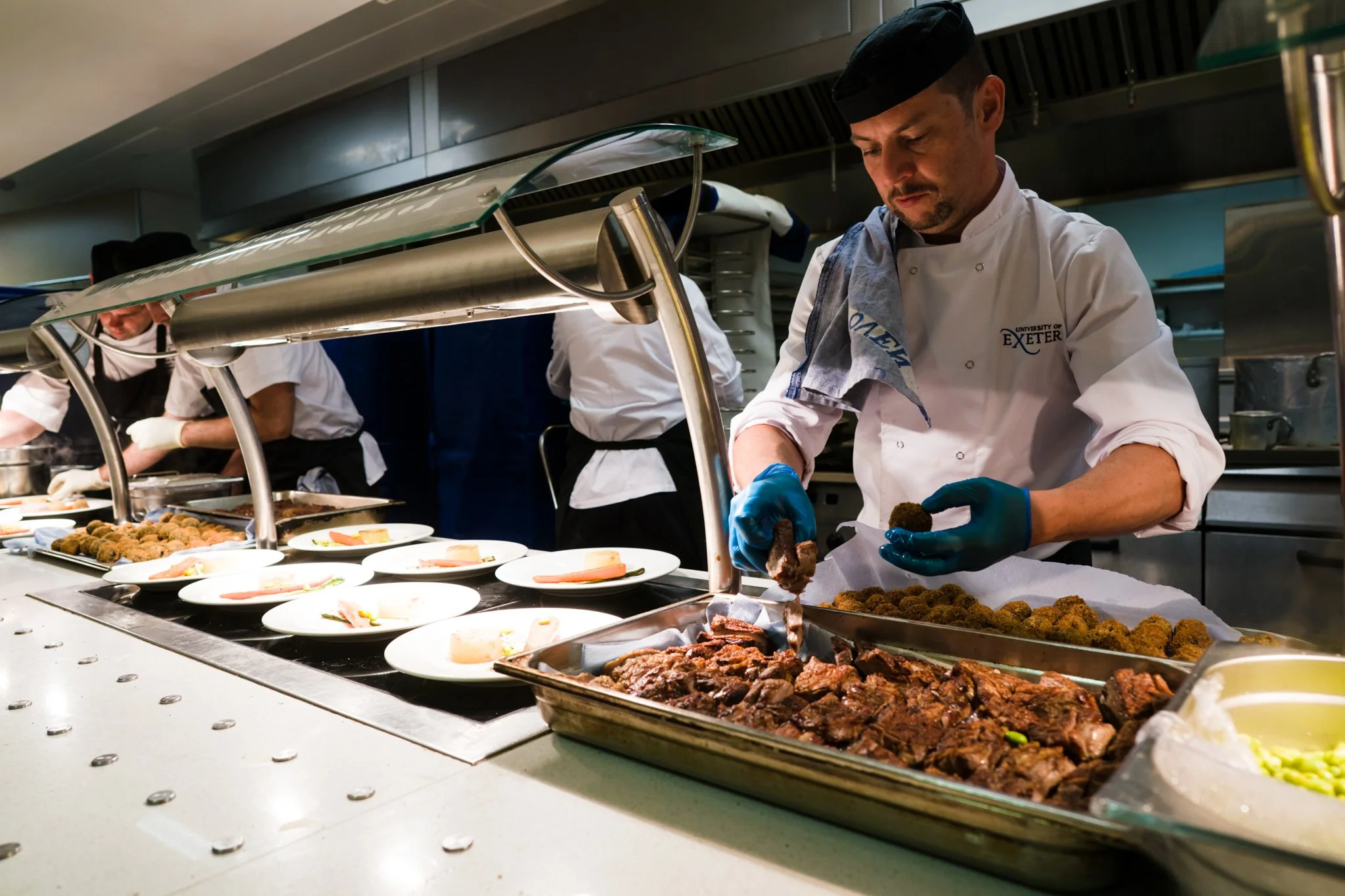 A chef in a white uniform and black hat preparing food in a professional kitchen, with multiple plates of food on the counter, including breaded items and sliced meat.