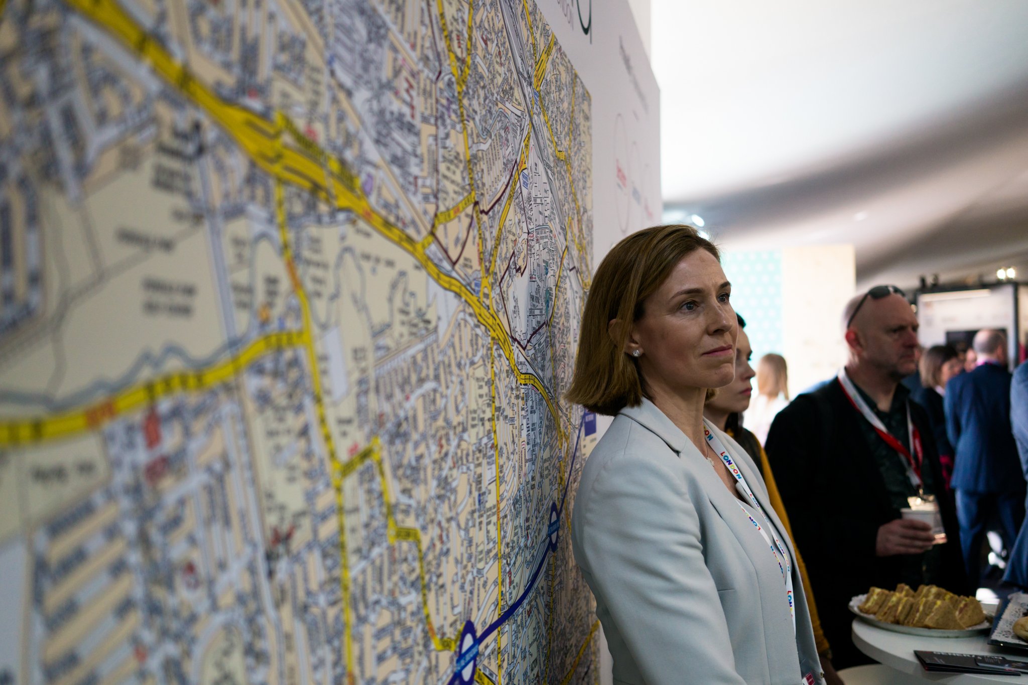 A woman with shoulder-length brown hair stands beside a large city street map, looking ahead thoughtfully, at a crowded indoor event.