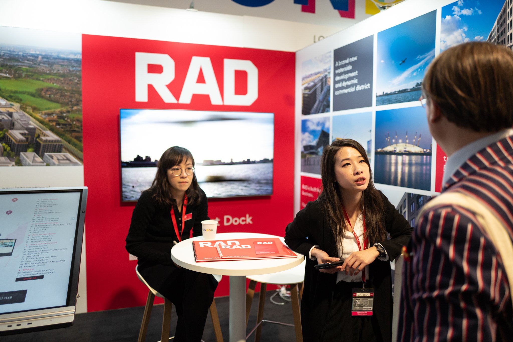 Three women are in conversation at an exhibition booth with a large red sign reading 'RAD'. One woman with glasses is sitting behind a round table with brochures, while the other woman with shoulder-length hair is standing and talking to a man with a