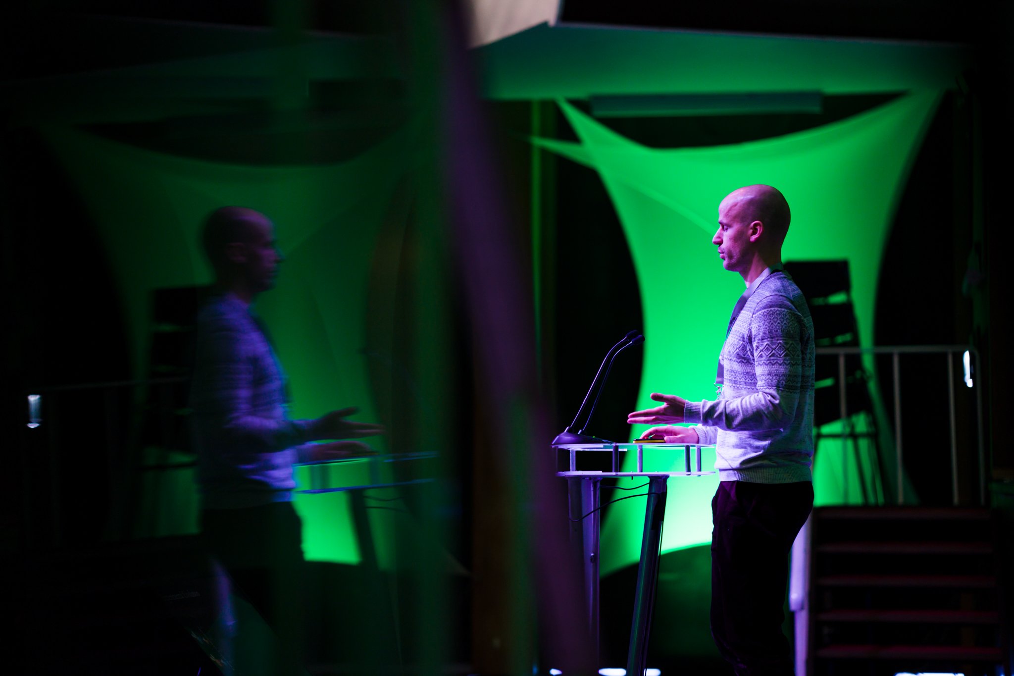 A man with a shaved head wearing a sweater and standing in front of a podium, speaking or presenting in a dimly lit room with green lighting and fabric decorations.