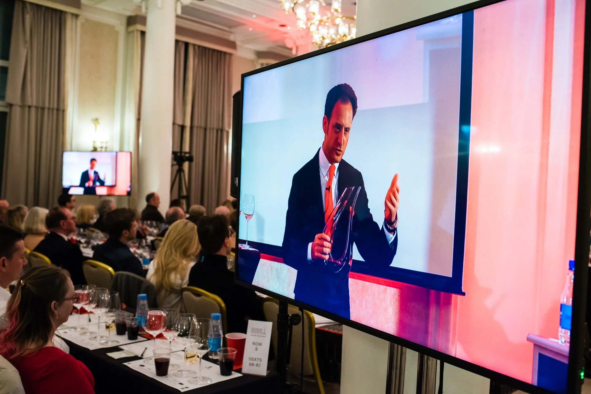 A man presenting at a conference with a large screen showing his image. The event has attendees seated at tables with glasses and bottles.