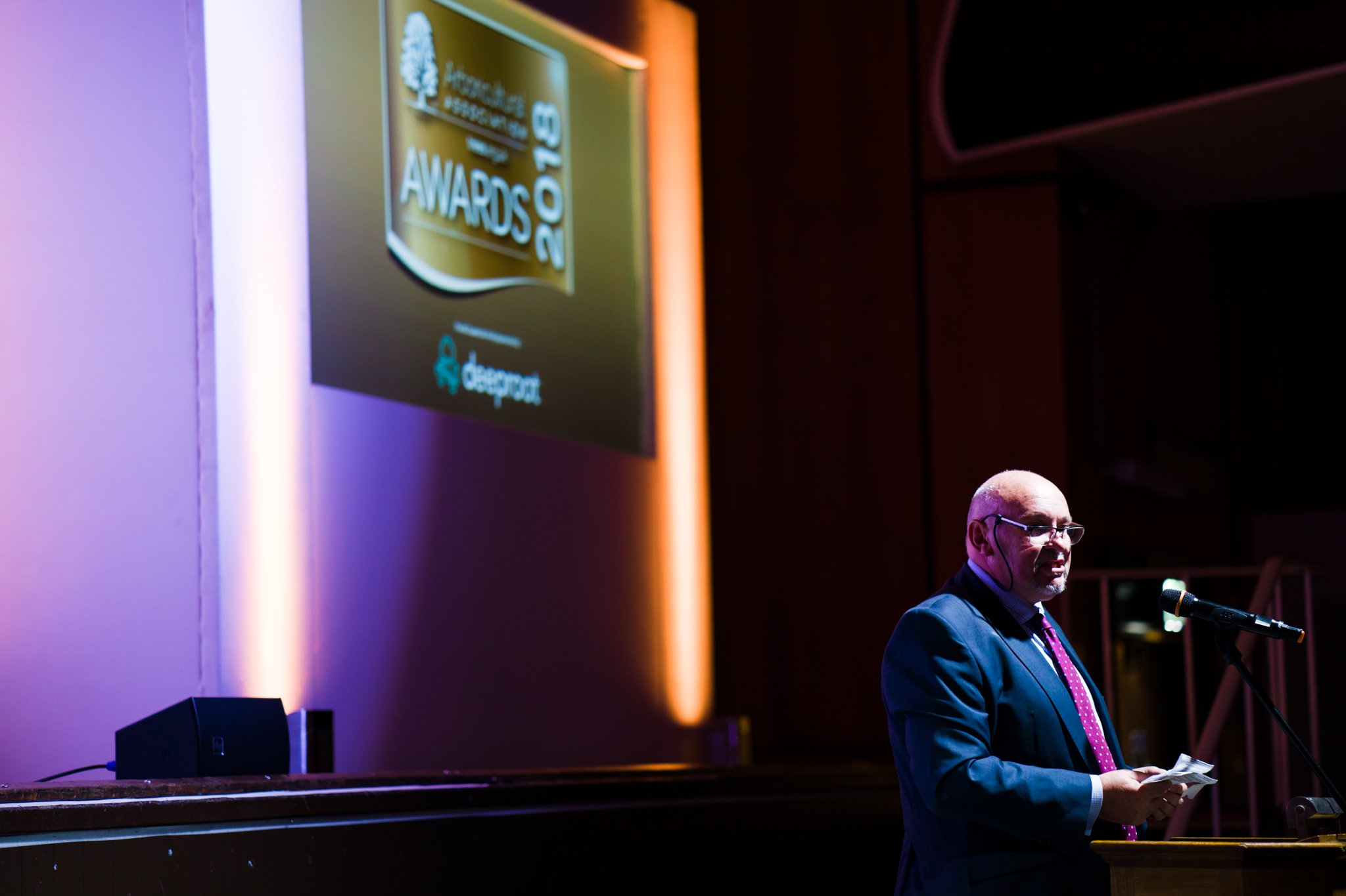 A man in a suit and pink tie speaking at a podium with a microphone at an awards ceremony, with a large screen behind him displaying the awards event logo.