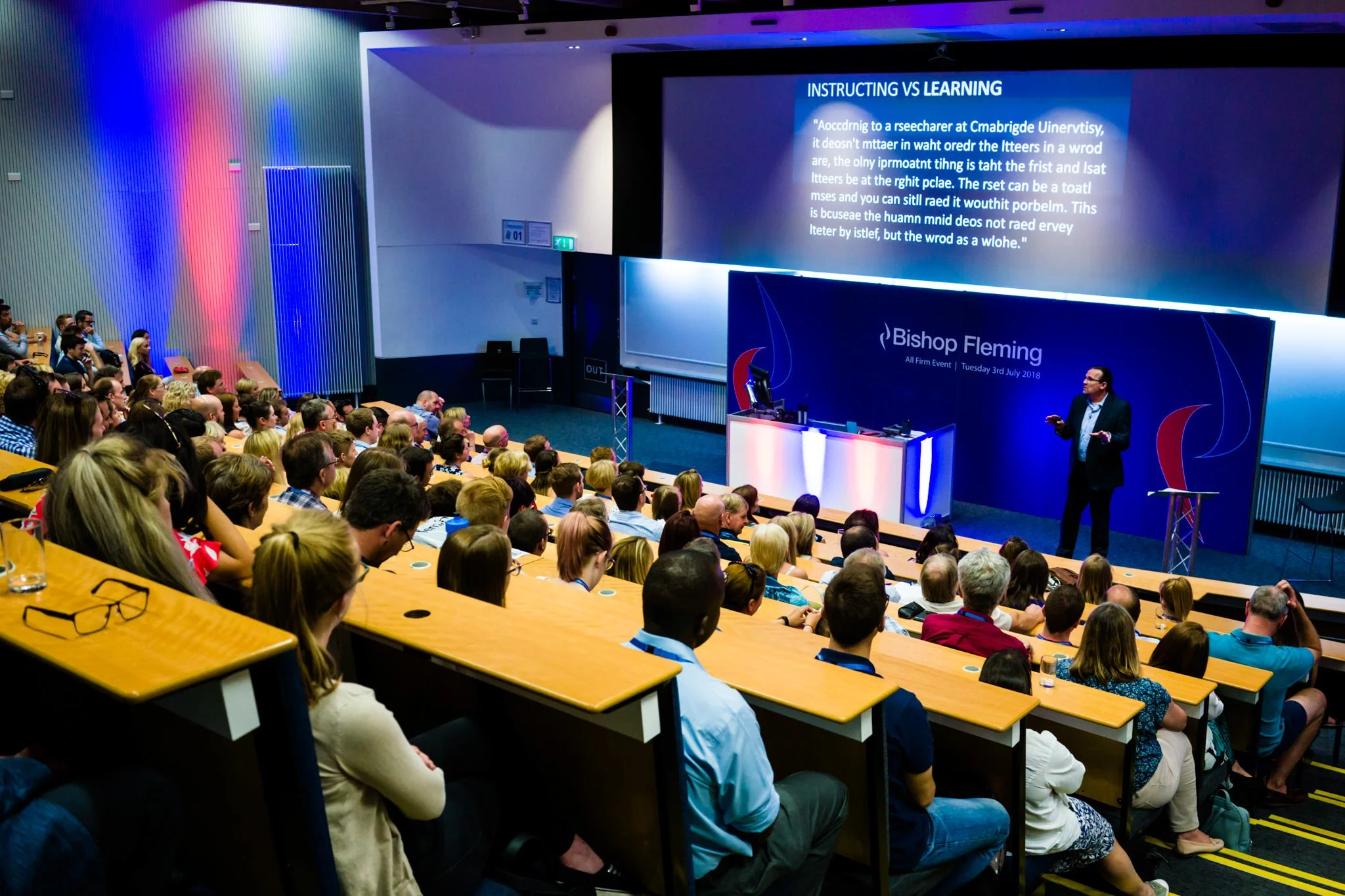 A large audience seated in tiered seats inside a conference hall listening to a speaker on stage. The stage features a large screen displaying a presentation and a sign that reads "Bishop Fleming". The audience appears engaged, with some taking notes