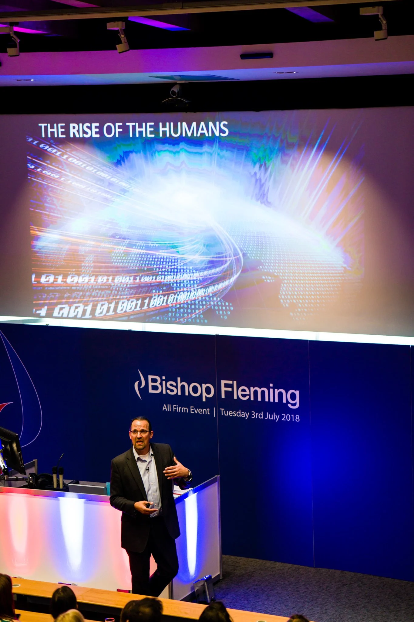 A man giving a presentation in a conference room with a slide titled 'The Rise of the Humans' projected behind him. The conference is hosted by Bishop Fleming on July 3rd, 2018.
