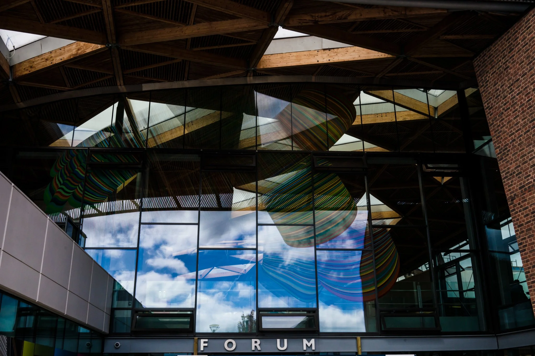 Reflection of clouds and sky in large glass windows of a building with an abstract colorful sculpture inside visible through the windows, and the word 'FORUM' on the exterior