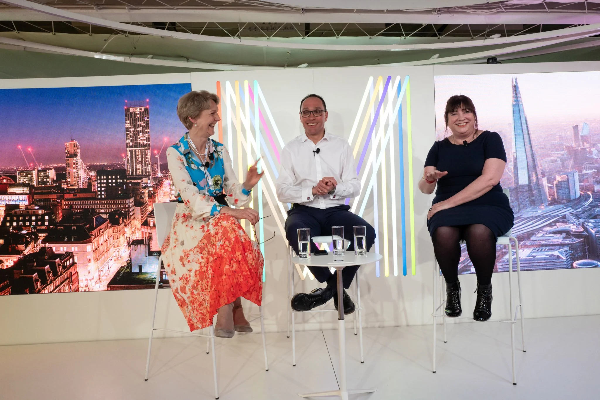Three people sitting on white chairs on a television set, with a backdrop of cityscape images and colorful vertical light strips, engaging in a lively discussion.