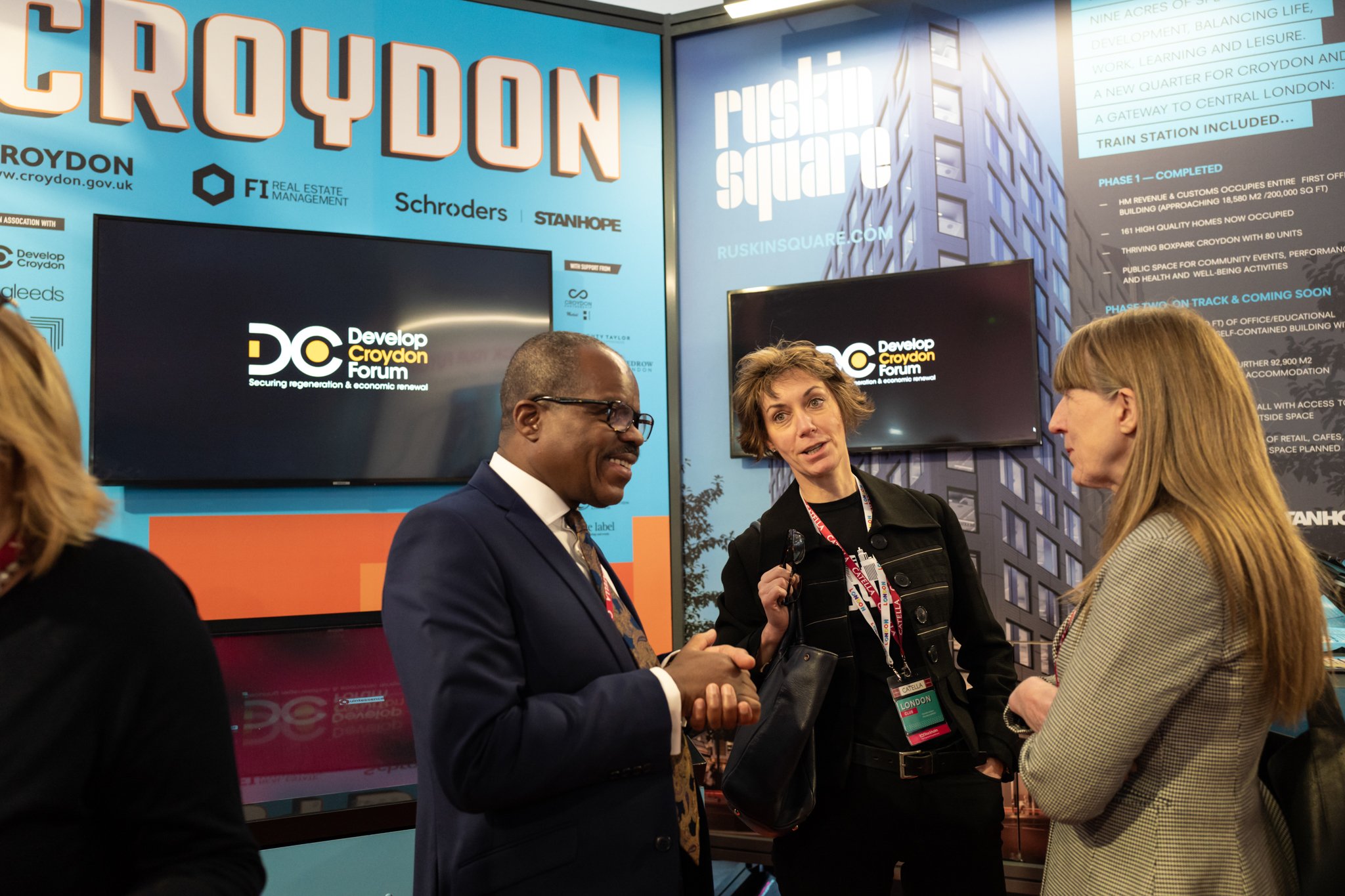 Three professionals engaged in conversation at the Develop Croydon Forum, with large screens and information about Croydon development projects in the background.