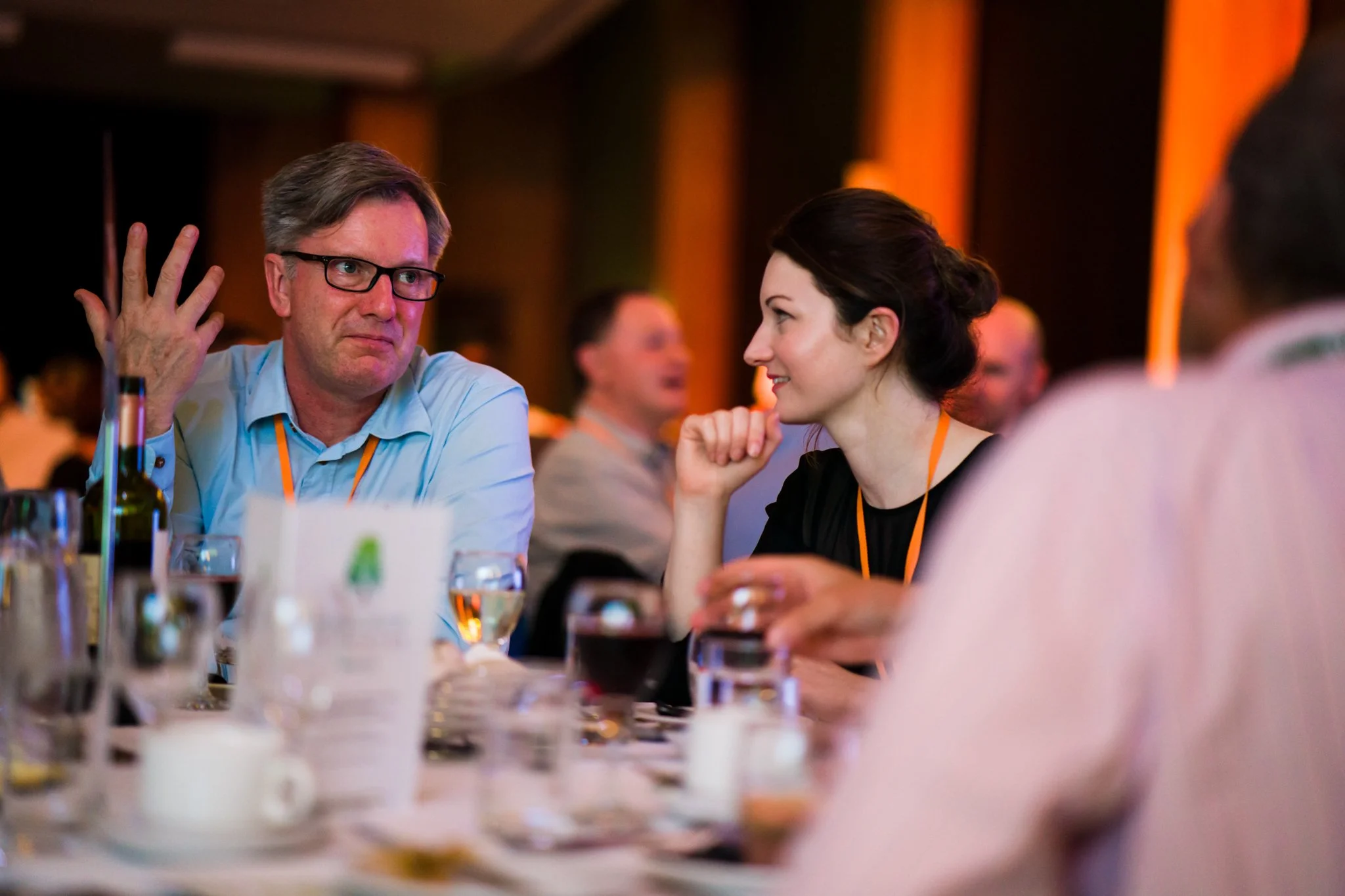 People at a dinner table engaging in conversation at an event or conference, with drinks and place settings on the table.