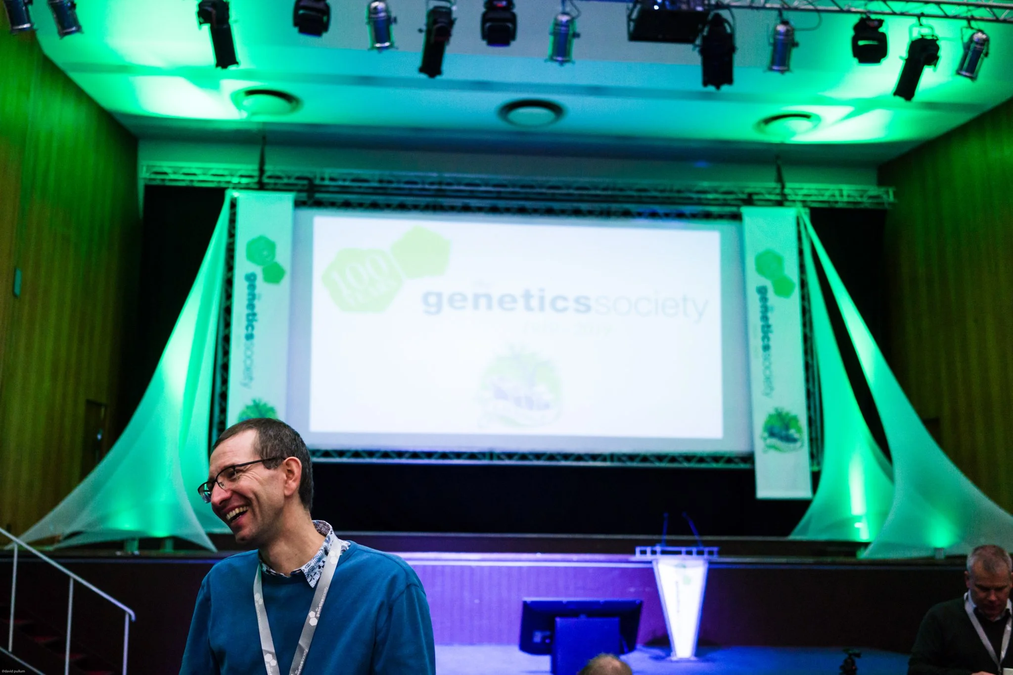 A conference room with a large screen displaying the logo for Genetics Society. The room is illuminated with green lighting, and there are people smiling and talking in the foreground.