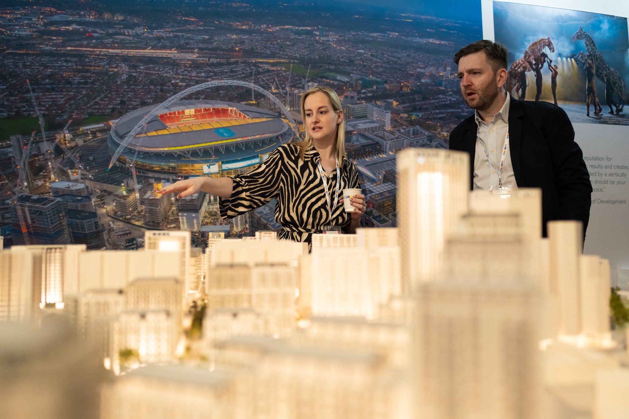 Two people discussing a scale model of a city, with a large aerial photograph of a stadium in the background.