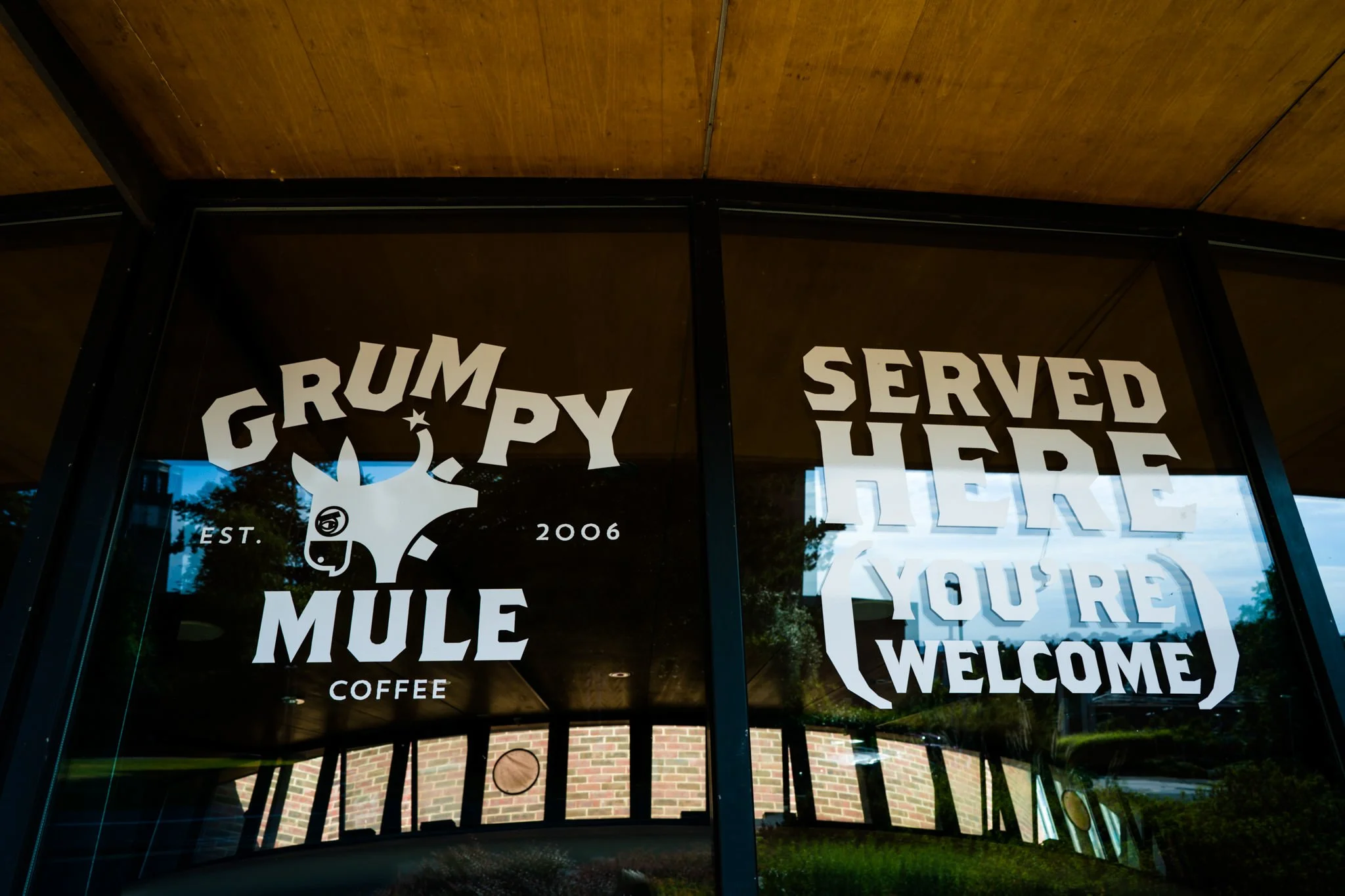 Sign on a window for Grumpy Mule Coffee, established in 2006, with a graphic of a donkey wearing glasses and a moon and star. The sign says 'Served Here' and 'You're Welcome'.