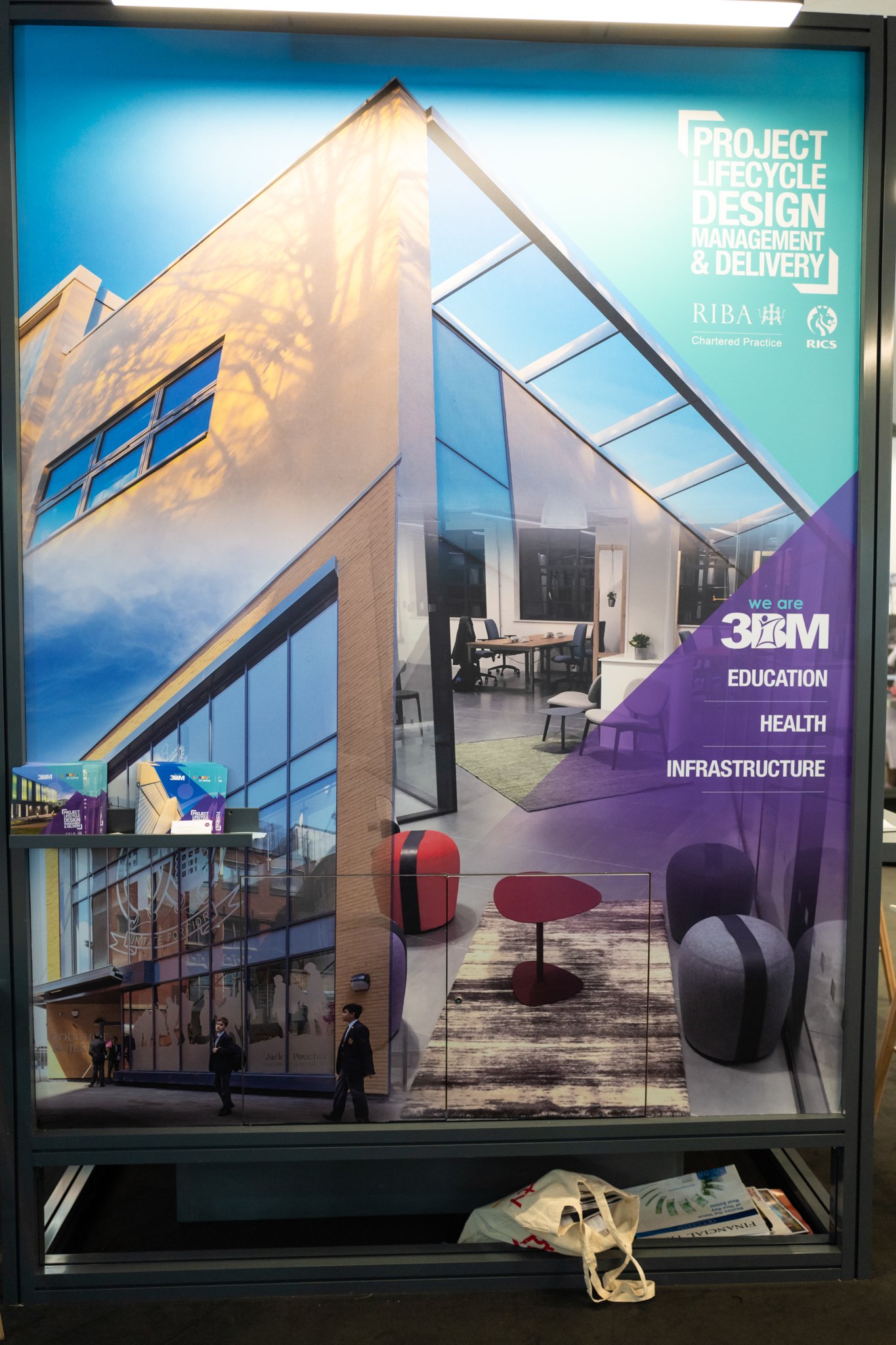 A promotional display poster showing a modern building with large windows, an interior space with seating and tables, and information about project lifecycle design management and delivery, including logos for RIBA and RICS. Text highlights education