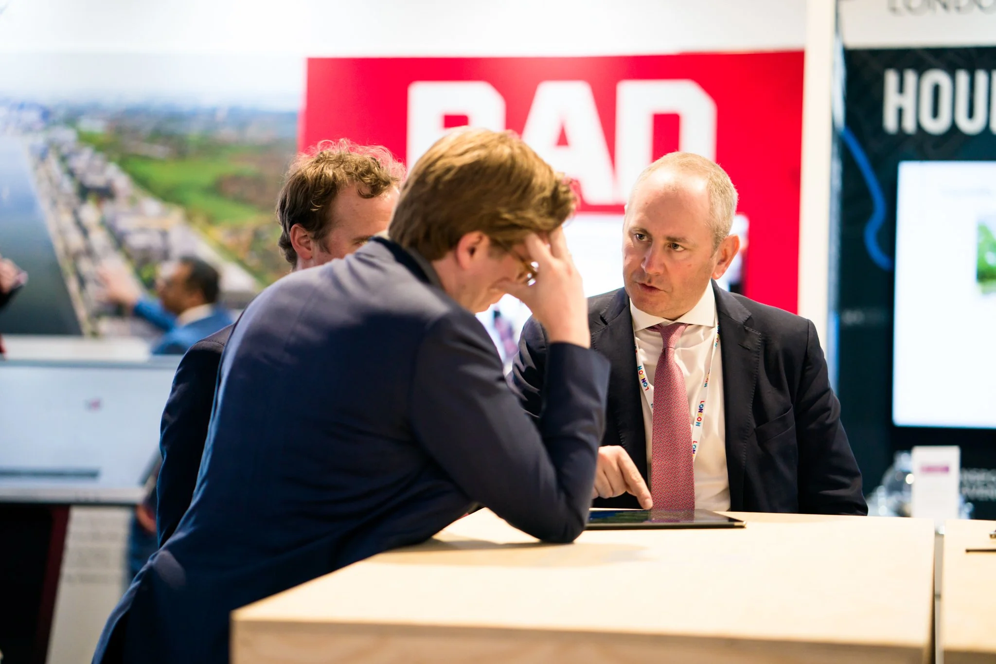 Three men in business attire having a serious conversation at a booth with a large red and white sign that says 'RAD' in the background.