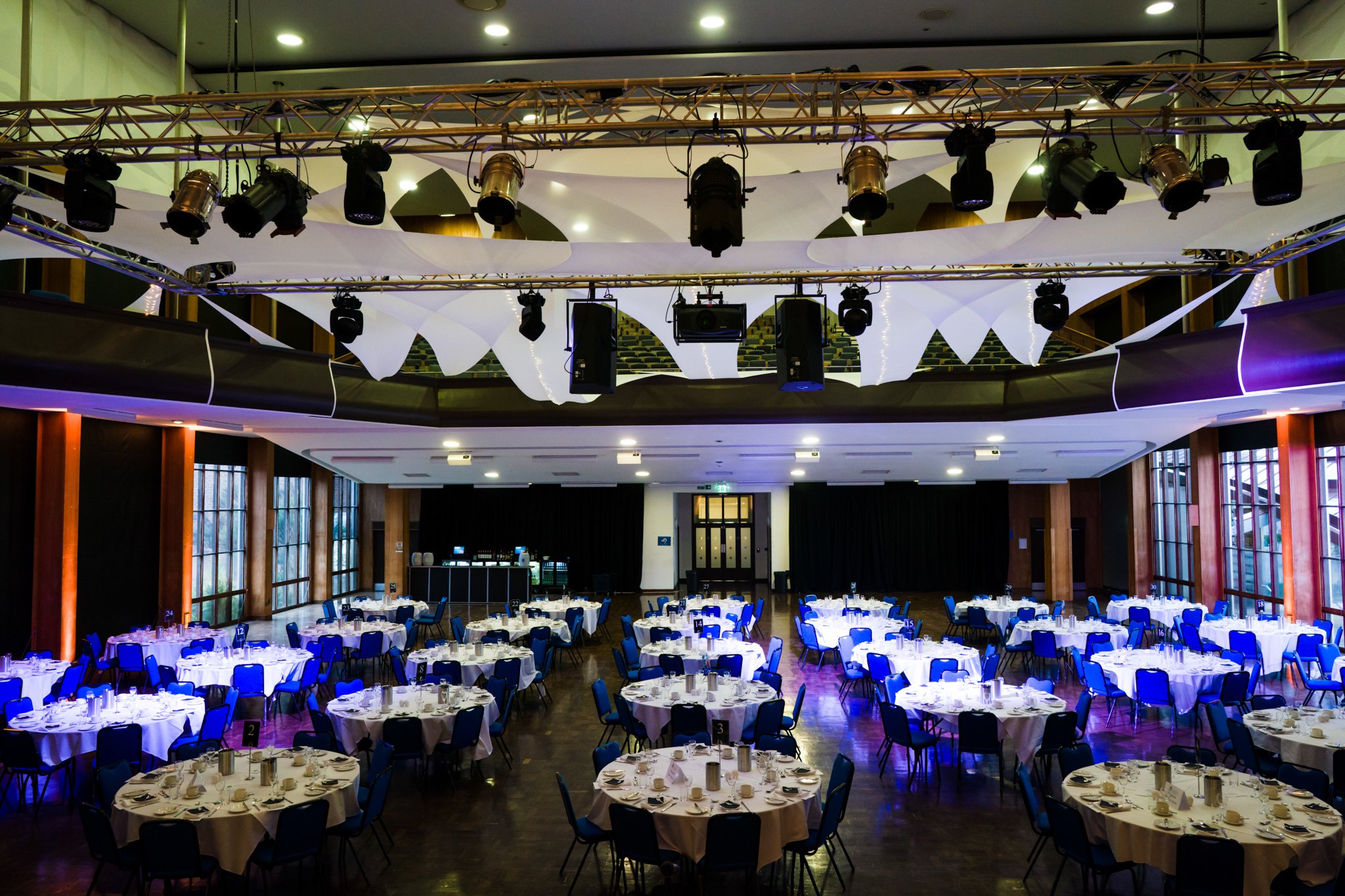 Undeveloped banquet hall with multiple round tables set for an event, surrounded by blue chairs, and a stage with lighting and sound equipment.