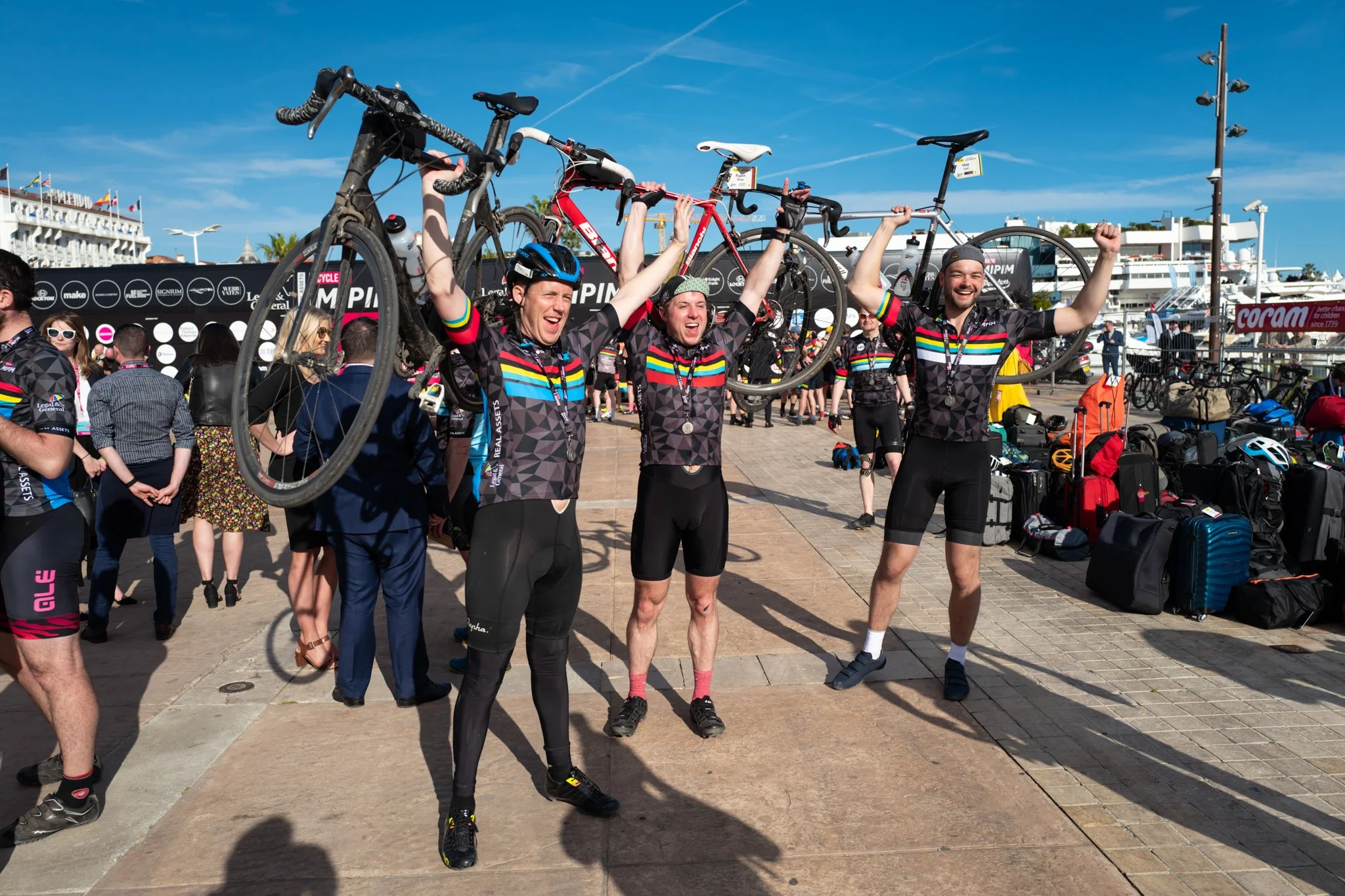 Cyclists celebrating with raised bicycles, wearing matching cycling jerseys, on a sunny day at a marina with bags and other cyclists in the background.