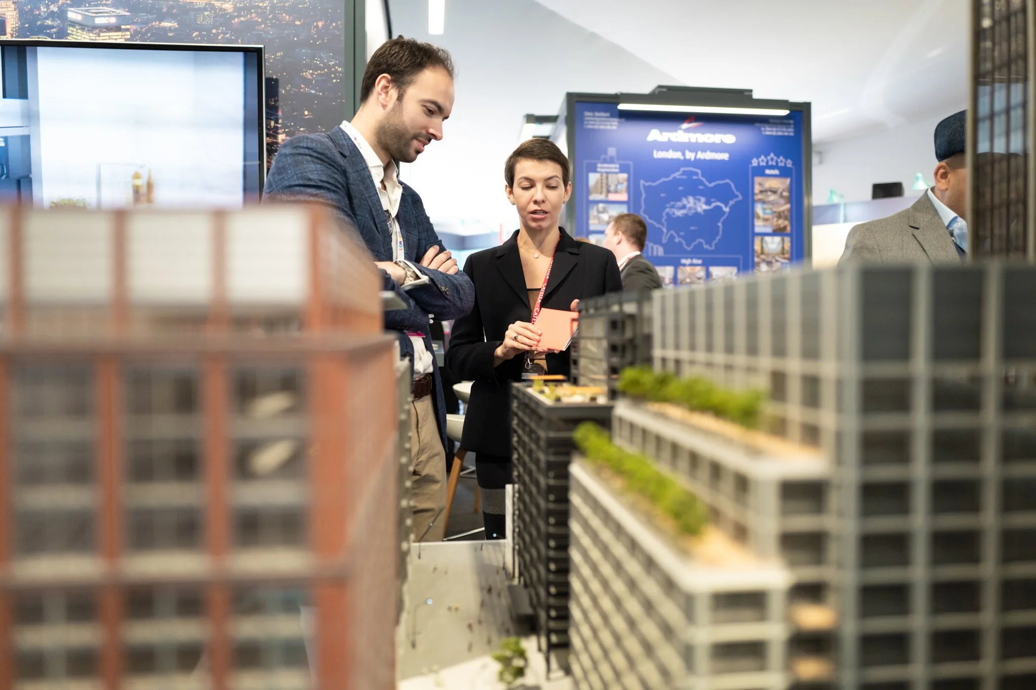 Two individuals examining a detailed architectural scale model of a urban development project at a professional event, with indoor setting and informational display in the background.
