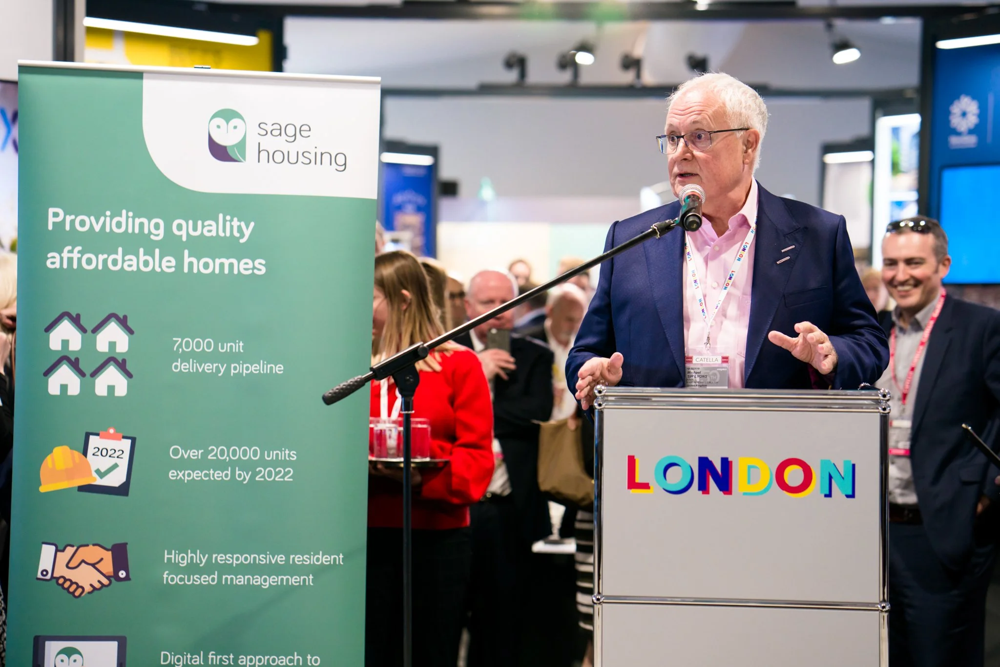 A man speaking at a podium with a sign that says 'LONDON' during an event. The speaker is wearing a navy suit, pink shirt, and glasses, with a lanyard around his neck. Behind him is a crowd of people, and a green banner promoting Sage Housing, highli