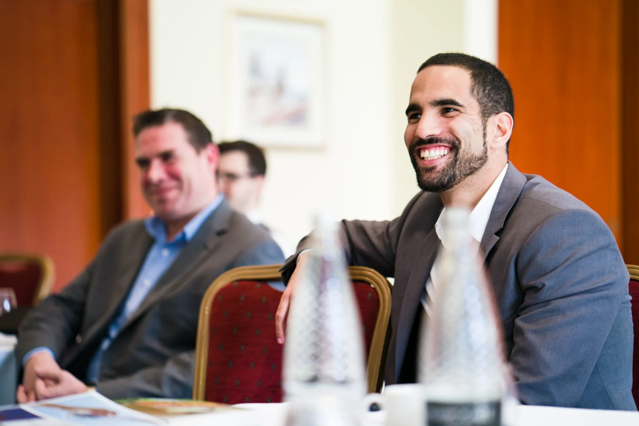 Two men in suits sitting at a conference table, smiling, with one prominently in focus in the foreground.