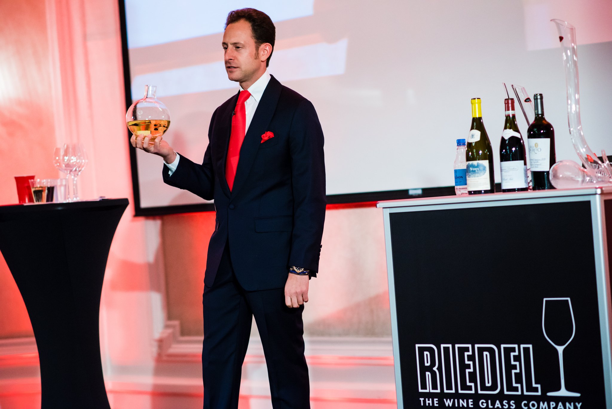 A man in a dark suit with a red tie and pocket square holding a glass decanter with a yellow liquid during a wine presentation or event. There are wine bottles and glassware on a bar counter next to a black sign with the logo 'RIEDL THE WINE GLASS CO