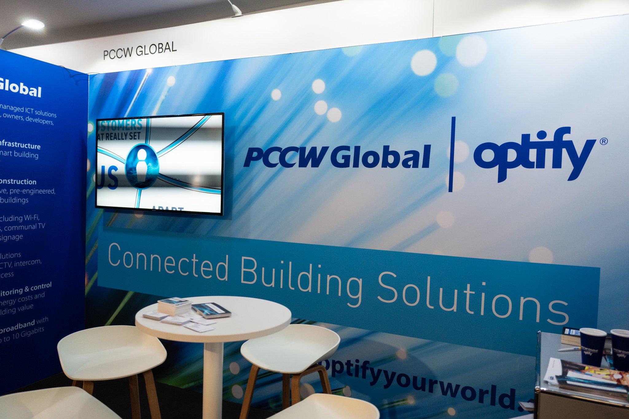 Exhibition booth for PCCW Global and Optify, featuring a blue background with company logos and the tagline "Connected Building Solutions." A TV screen displays information, and there are white chairs and a table with brochures and cups.