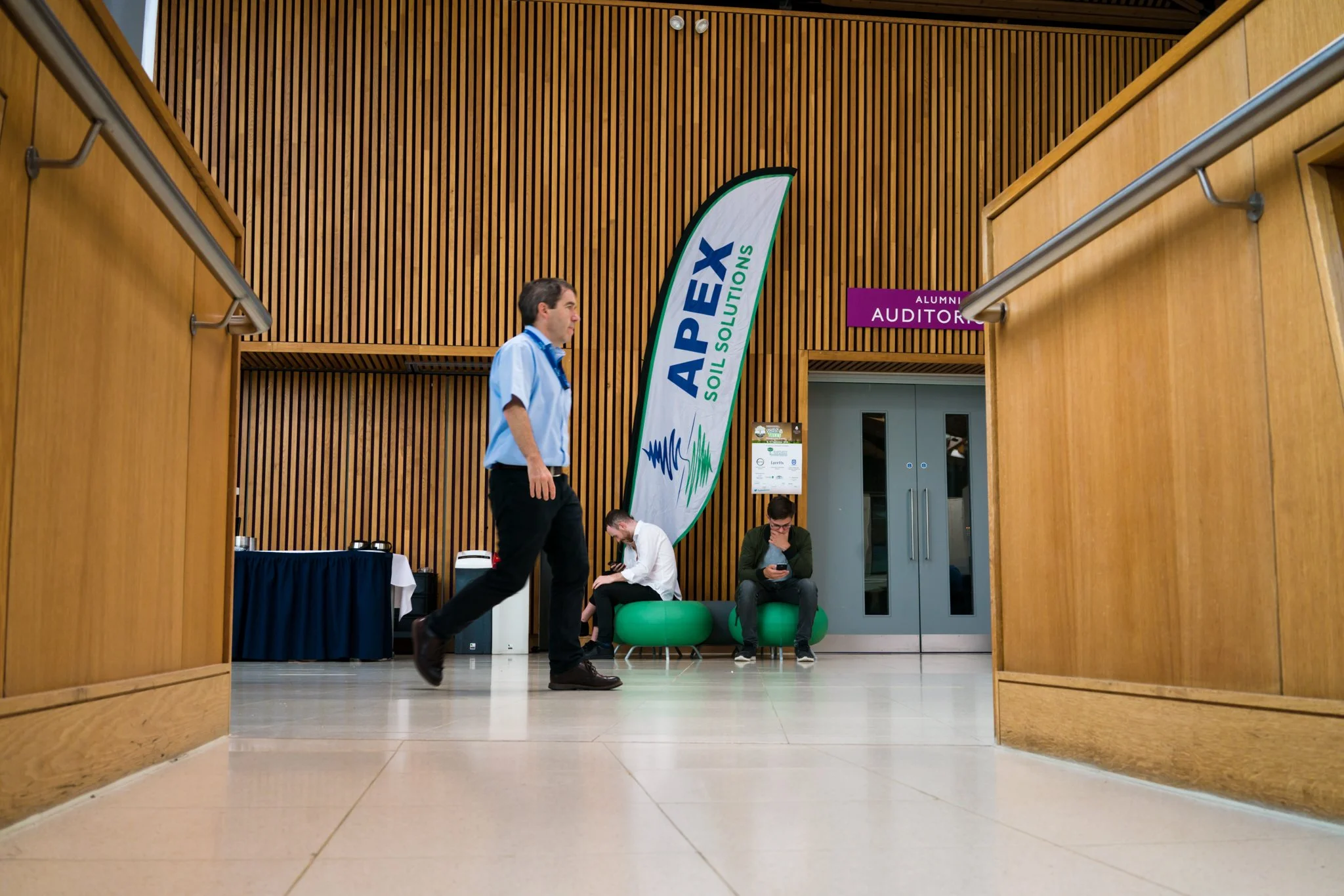 Interior of a conference center or auditorium with wooden walls, a large vertical banner for 'APEX SOIL SOLUTIONS,' and people sitting on green benches, one person walking in the foreground.