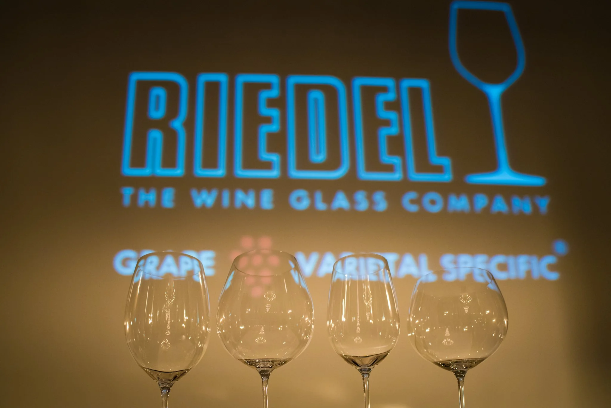 Four empty wine glasses in front of a backlit sign that reads "BUEDEL THE WINE GLASS COMPANY" with a wine glass icon, and the additional text "GRAPE VARIETAL SPECIFIC"