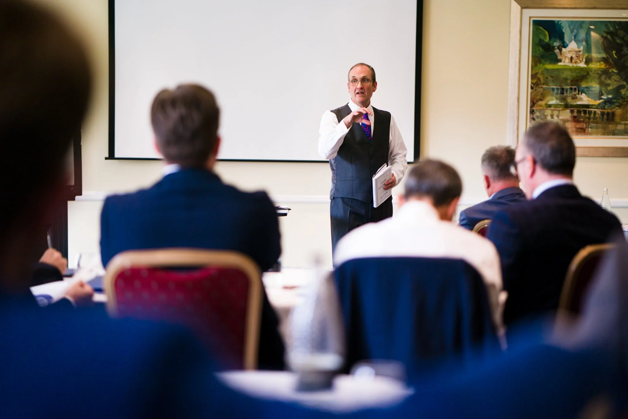 Man in formal attire giving a presentation to a group of business professionals in a conference room.