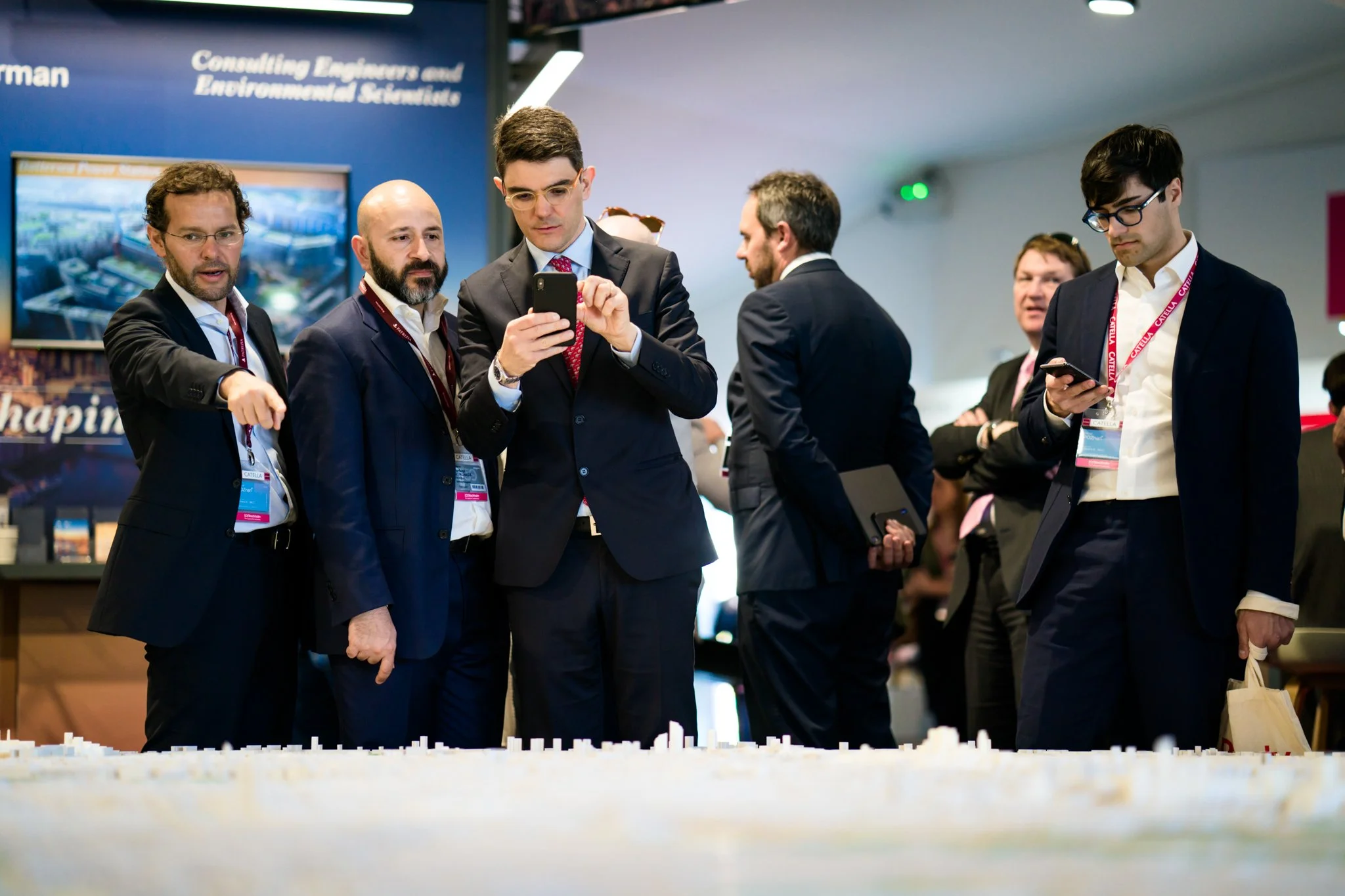Group of professionals in business suits at a conference, with some looking at their phones and one pointing at a large model or display on a table.