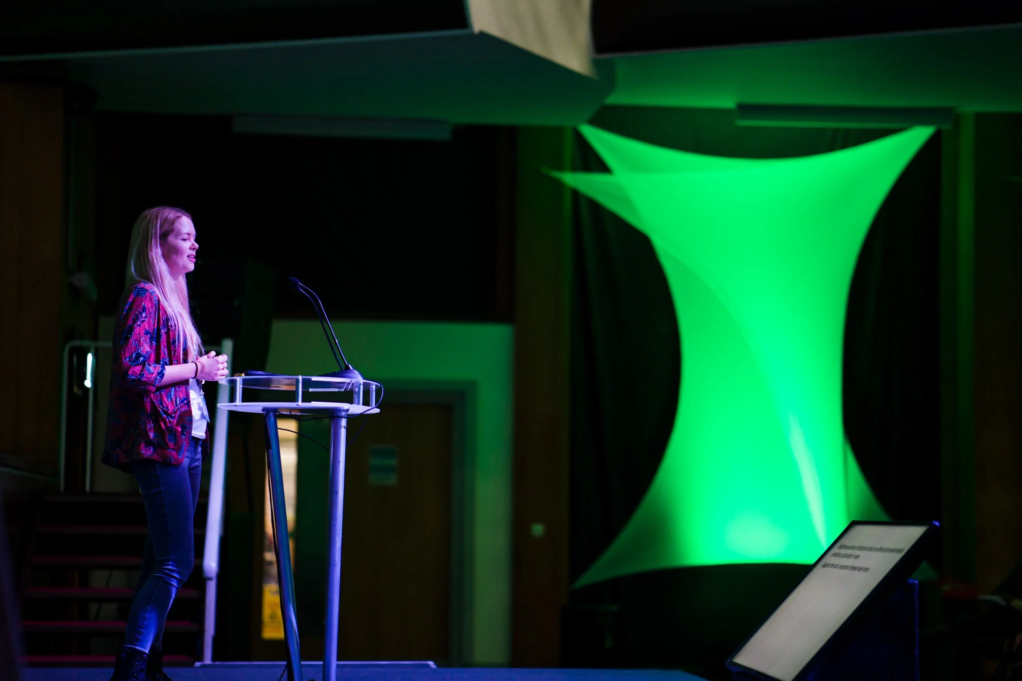 A woman giving a presentation on stage, standing beside a transparent podium with a microphone, in front of green stage lighting and a colorful backdrop.