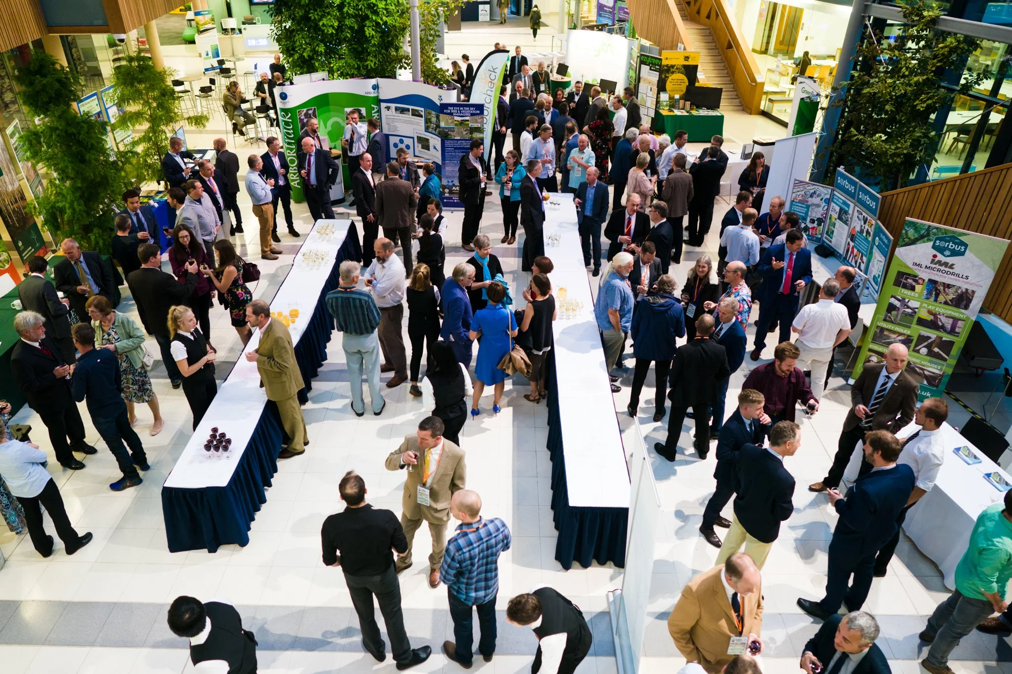 A busy conference or networking event with many attendees mingling, talking, and walking around displayed booths and tables with informational posters and materials inside a large indoor space.