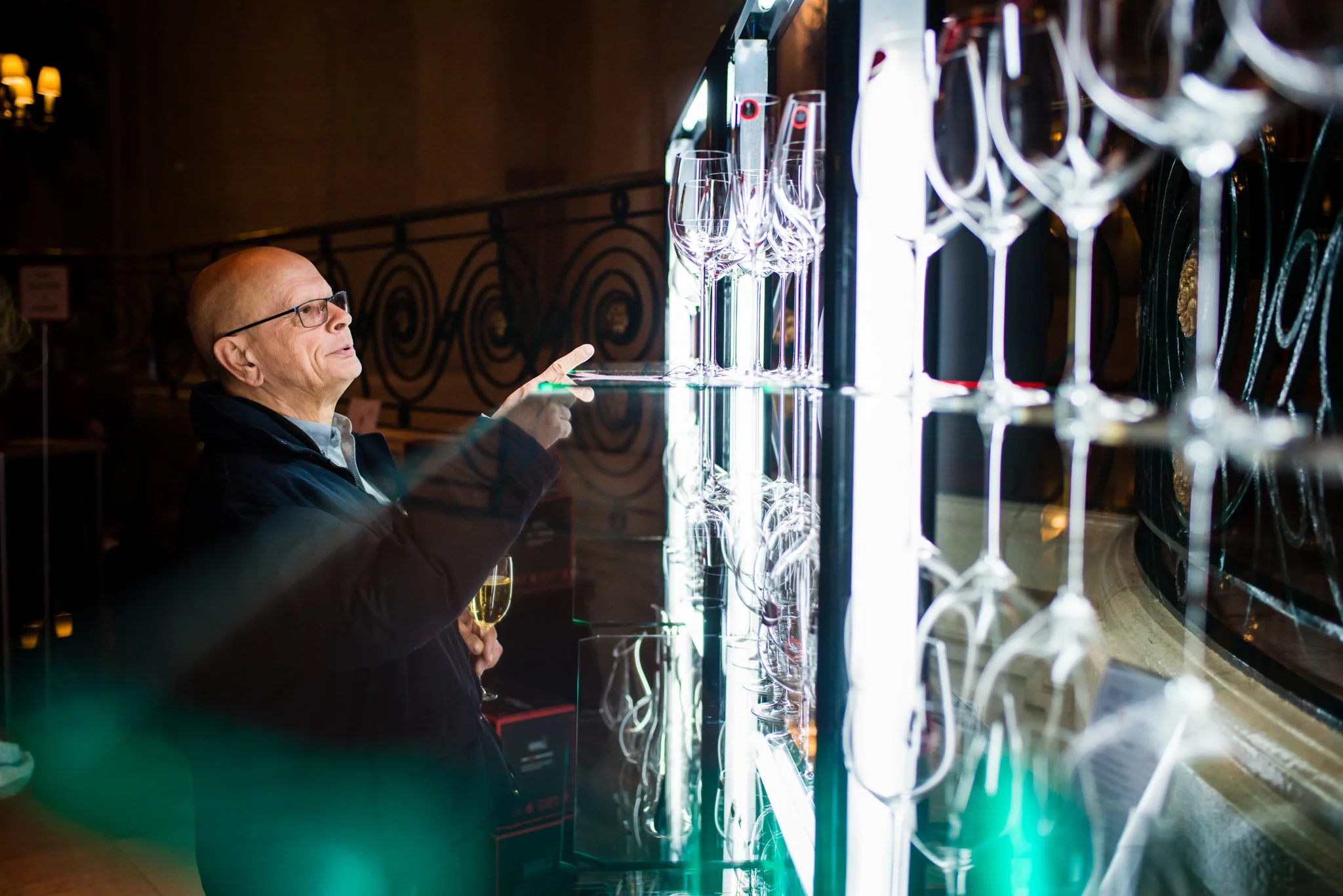 An elderly man with glasses holding a glass of champagne, pointing at crystals on display in a store or exhibition.