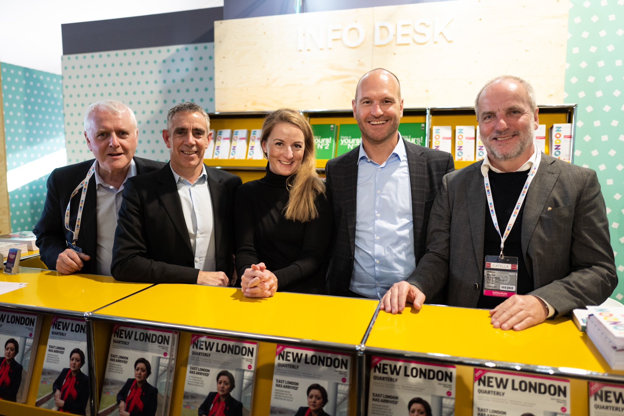 Five people standing behind a yellow counter with magazines on it at an event, with a sign that says 'INFO DESK' in the background.