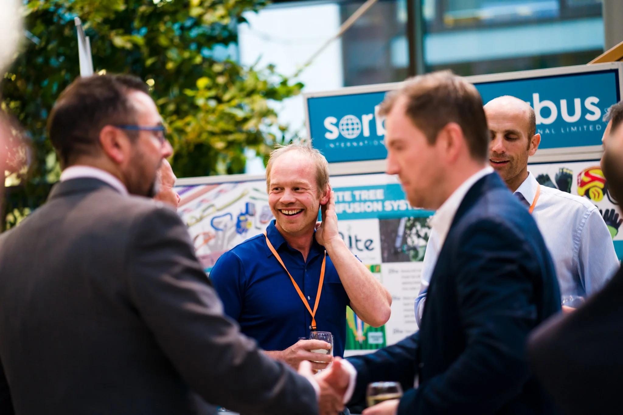 A group of men in business attire socializing at an outdoor event, with one man in a blue shirt smiling and touching his ear, while others are engaged in conversation and holding drinks.