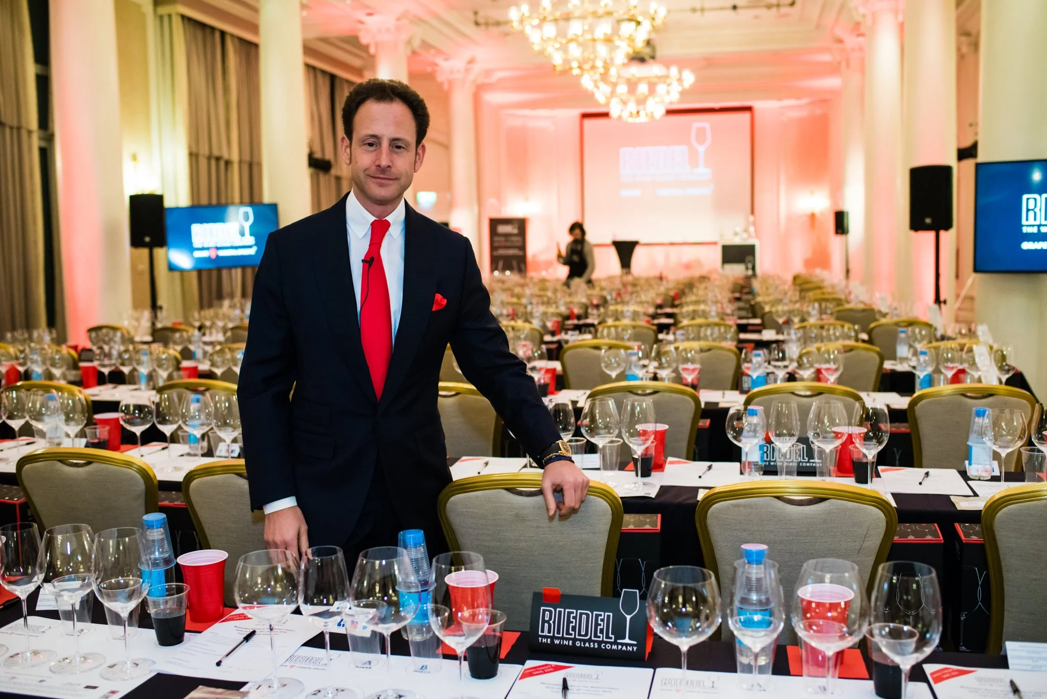 A man in a black suit with a red tie and pocket square standing in a large, decorated conference room with many tables set for a wine tasting event. The room has chandeliers, pink lighting, and presentation screens displaying the logo of Riedel, The 