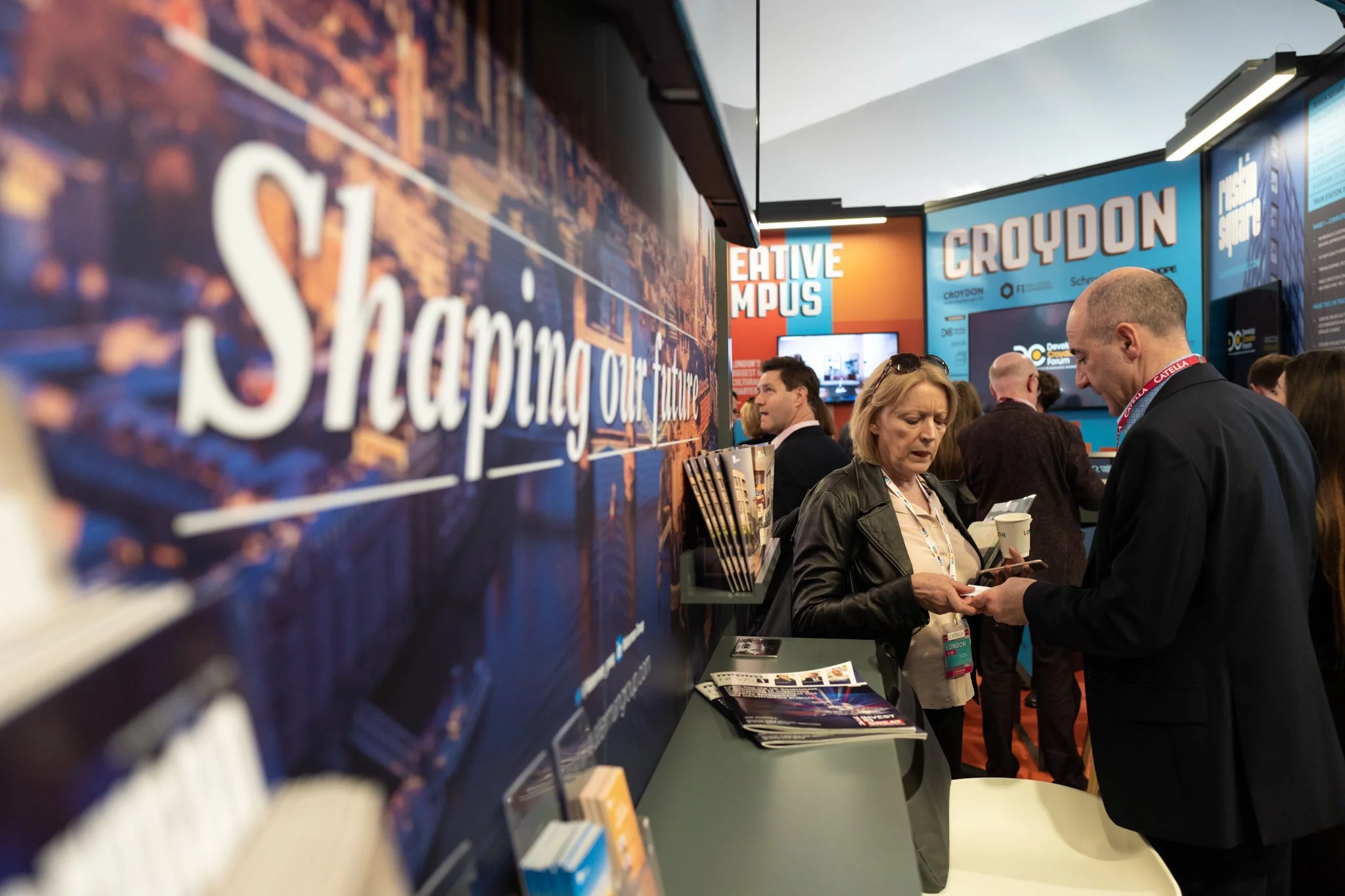 People at an indoor event, engaged in conversation near a booth with signs reading 'CREATIVE CAMPUS' and 'CROYDON.'