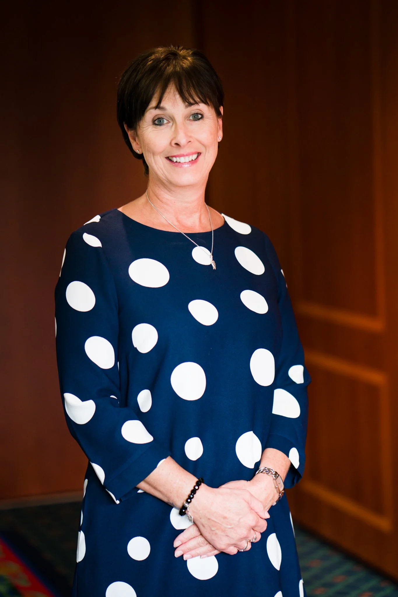 A smiling woman with short dark hair wearing a navy blue dress with white polka dots, standing in front of a wooden background.