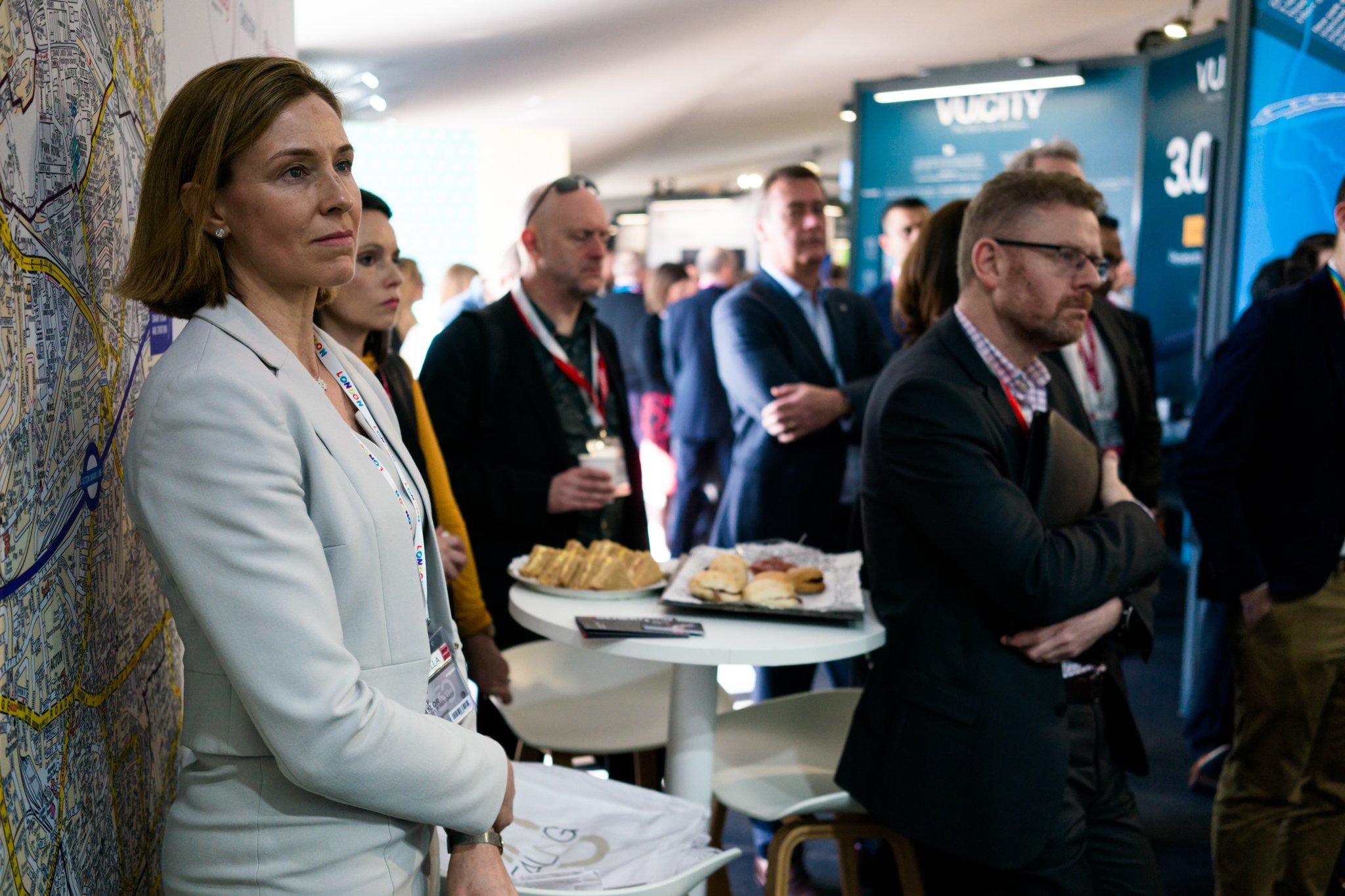 A group of people attending a conference or event, with some standing and listening attentively. One woman in a white blazer is prominent in the foreground, while others are in the background near a blue booth and food table with sandwiches.