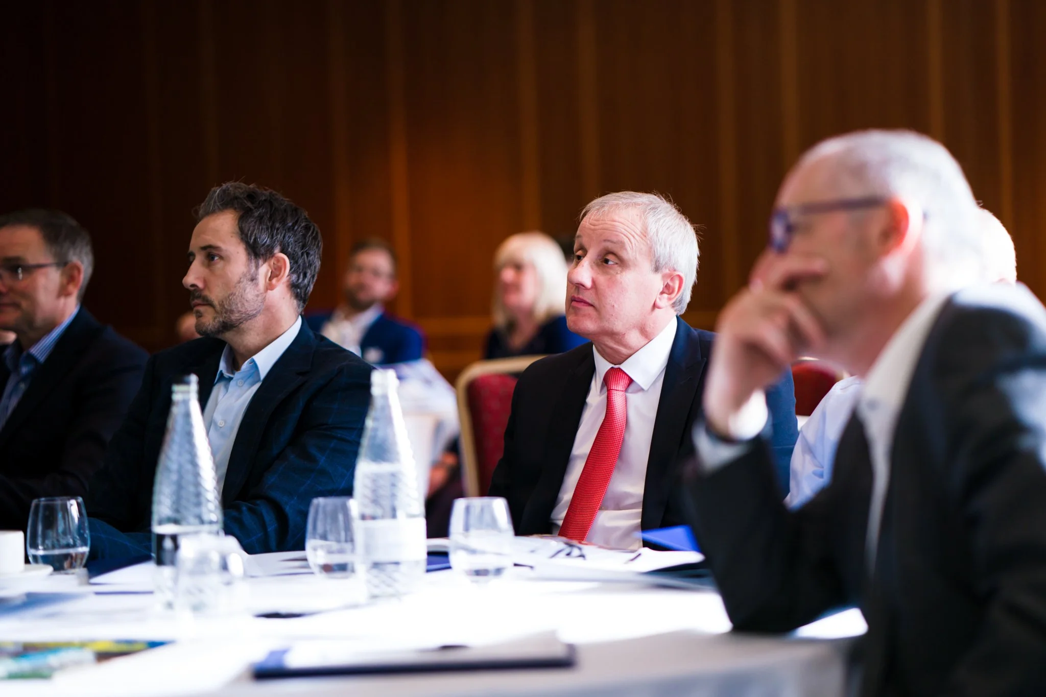 Business professionals attending a conference or meeting, sitting at a table with water bottles and glasses, listening attentively.