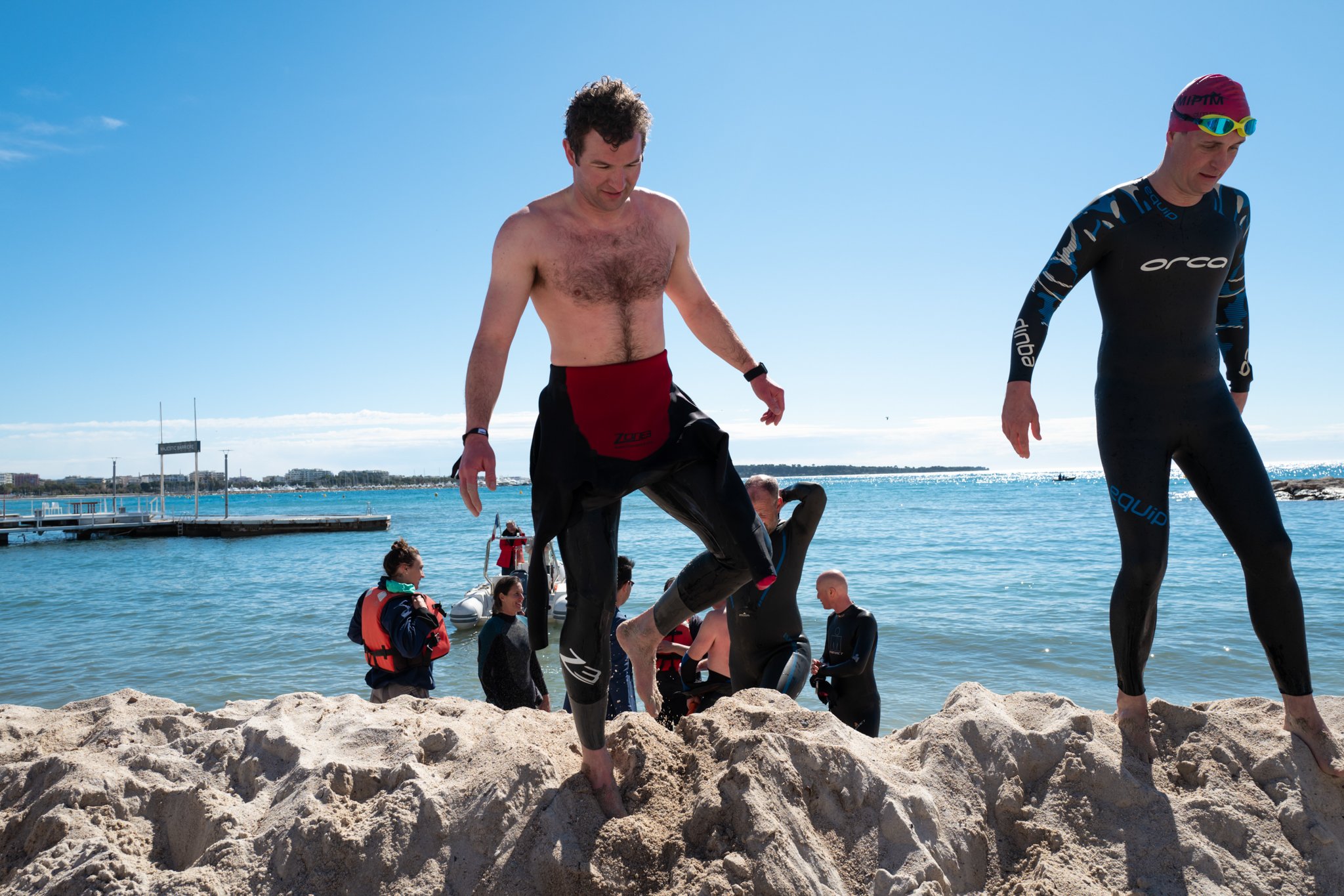 People arriving at a beach, some wearing wetsuits, with a pier and boats in the water, and a clear blue sky.