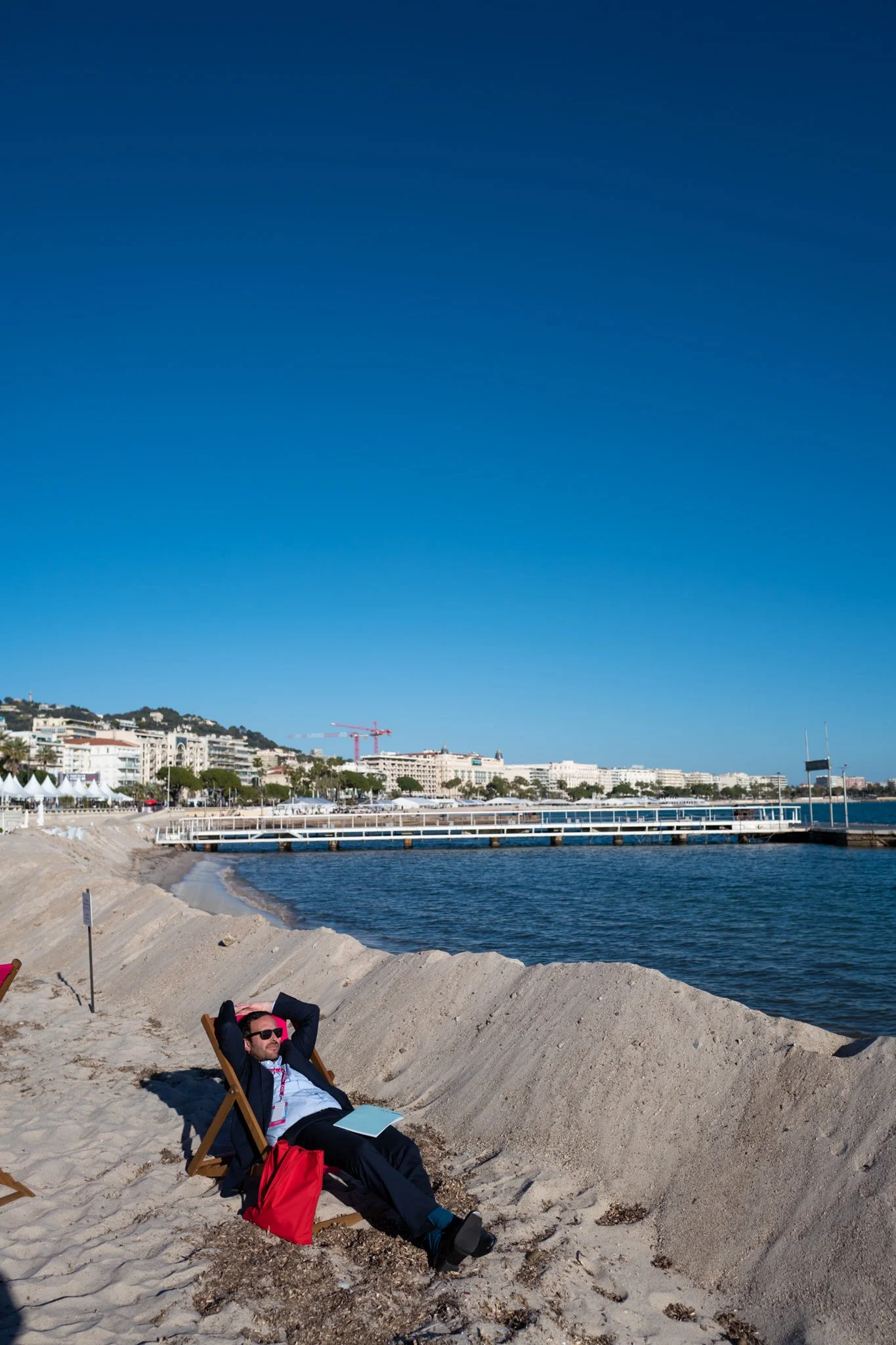 A man in business attire relaxing on a beach chair by the water, with a cityscape and clear blue sky in the background.