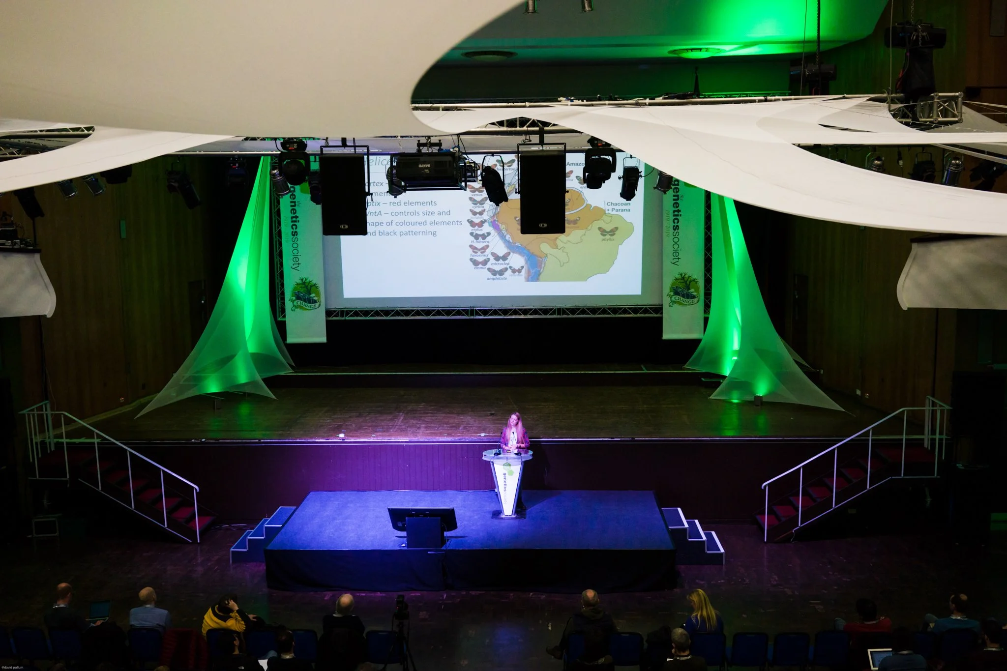 A woman is on stage giving a presentation with a large screen behind her displaying a slide about genetics, featuring diagrams and text. The stage is decorated with green lighting and fabric drapes, with an audience seated in front.