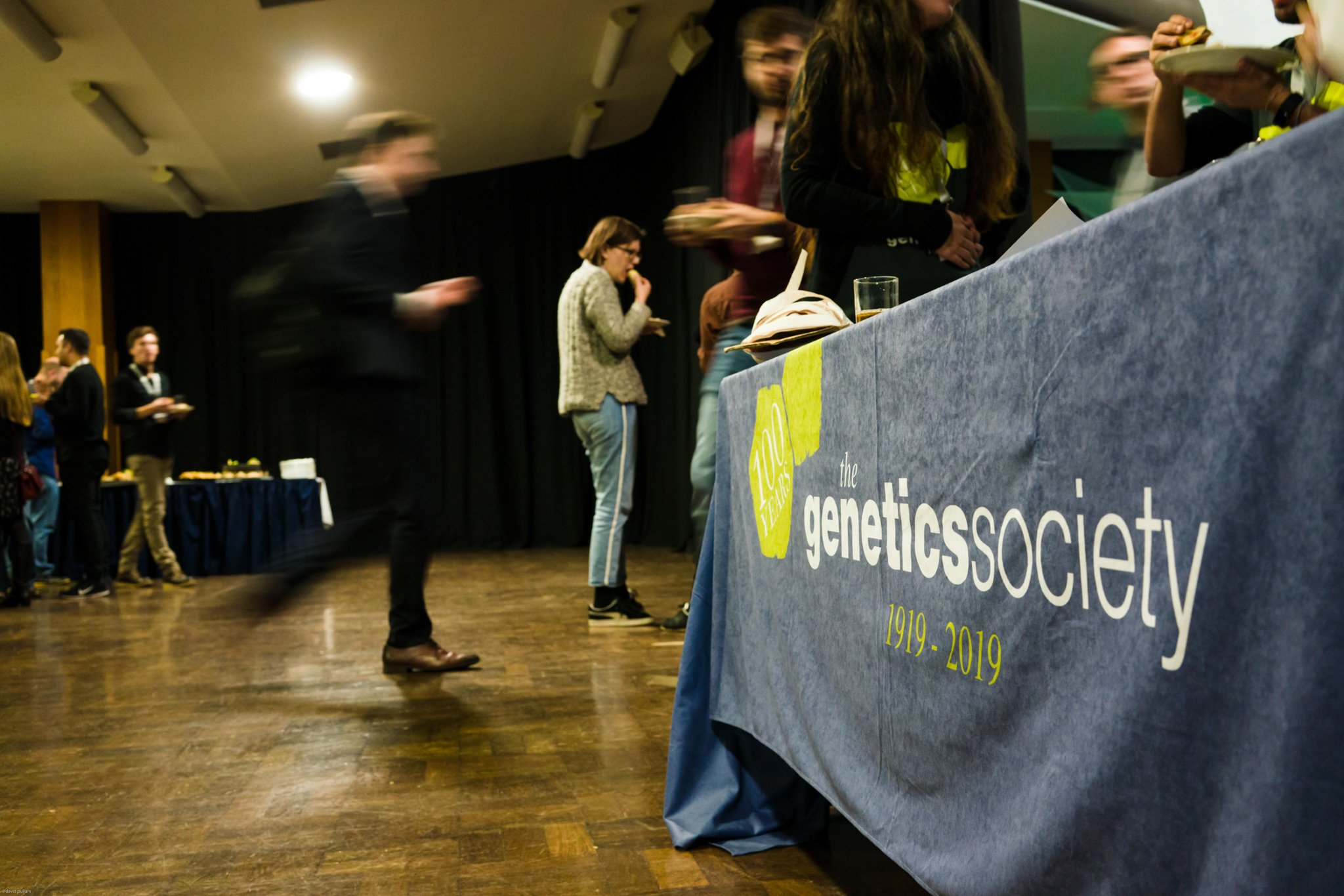 A group of people at a genetics society event, with some standing in line for food near a table with a blue tablecloth that reads 'the genetic society 1919-2019'. The room has wooden flooring and black curtains in the background.