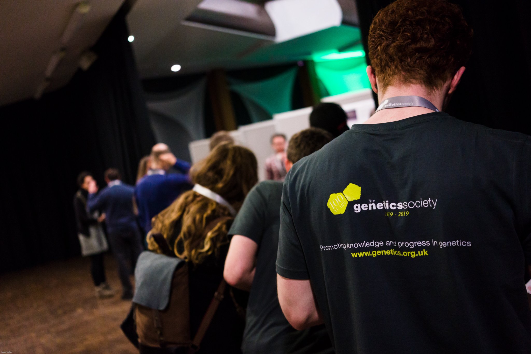 People attending a genetic society event, with a person in the foreground wearing a black T-shirt with the society's logo and website, and others in the background waiting or talking.