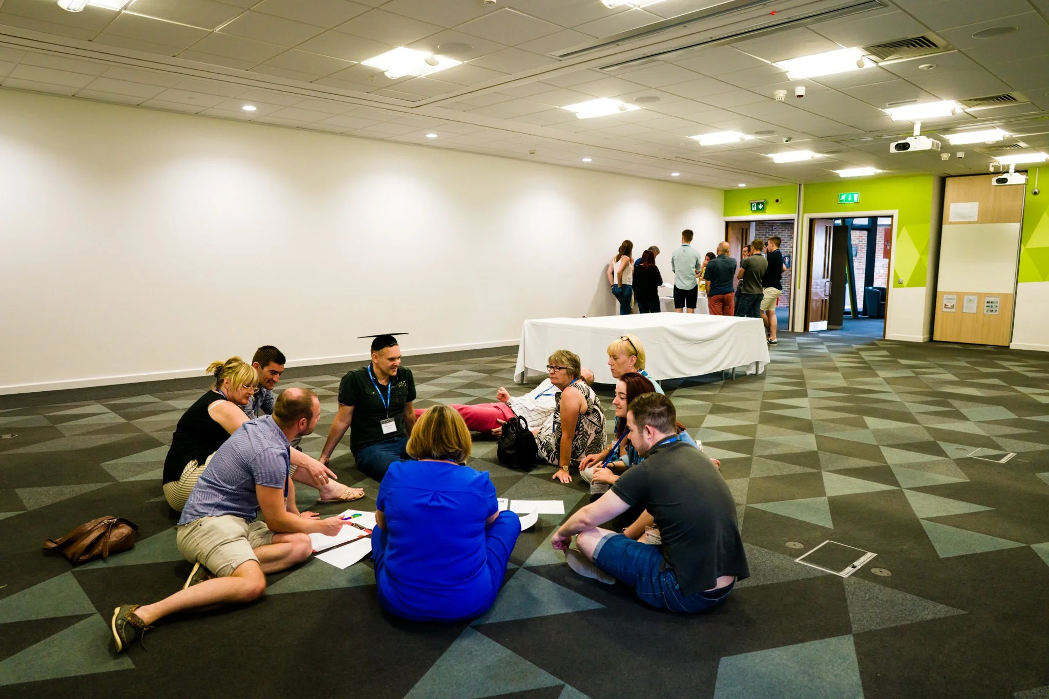 Group of people sitting in a circle on a patterned carpet in a conference room, with a few more people gathered near the door in the background.